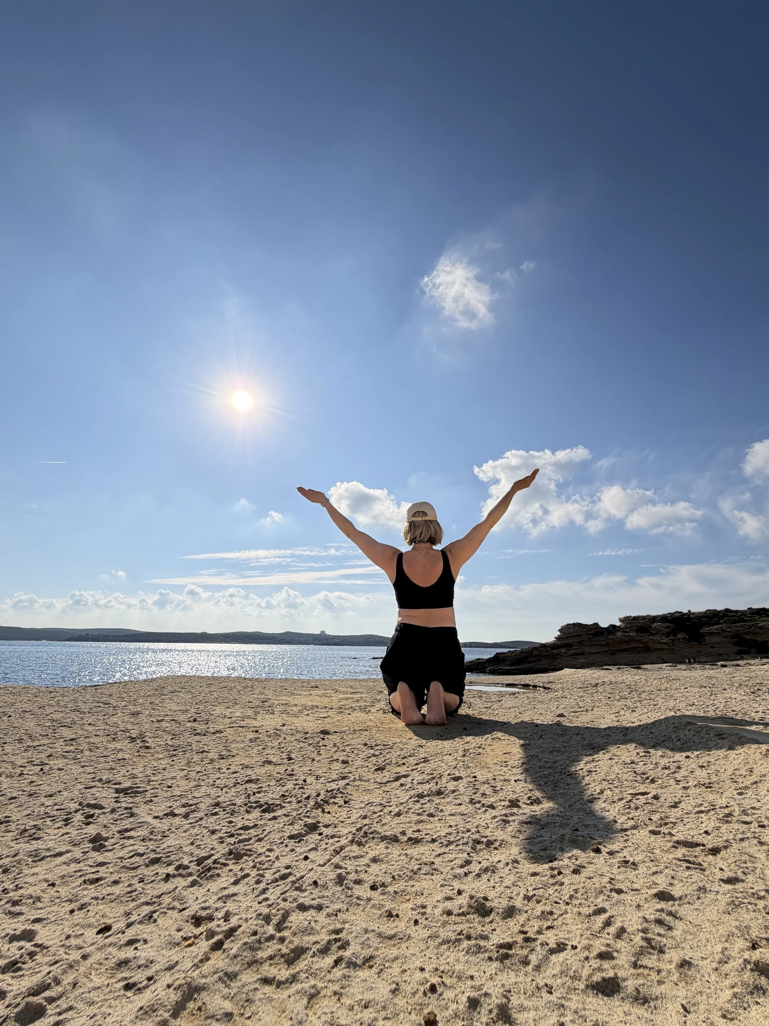 Woman sits with her arms in the air, back to camera. Sits on a sandy beach looking out to sea with a blue sky above