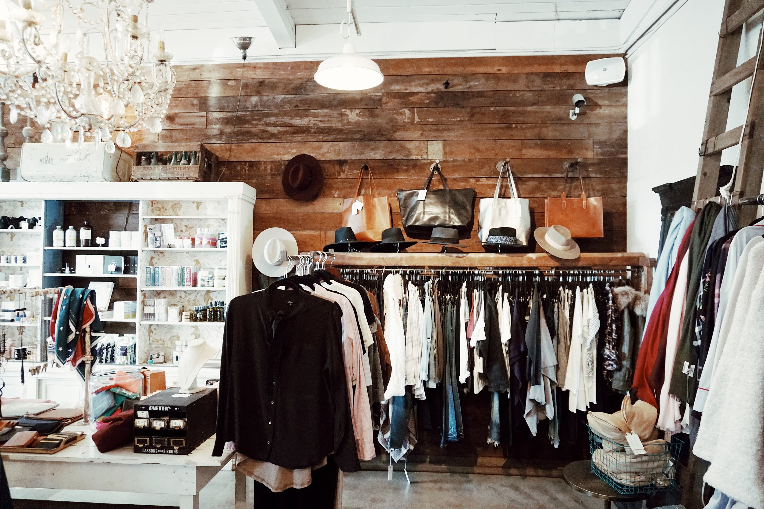 A cozy boutique interior featuring a rustic wooden wall with hats on display, chic Nashville-inspired handbags on a shelf, and a variety of clothes hanging from a metal rail along with accessories on a table.