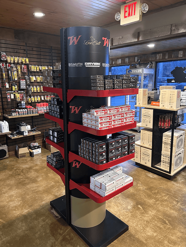 A retail display stand with multiple shelves stocked with Milwaukee tool accessories, located inside a store with various products and an exit sign visible.