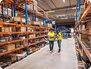 Two warehouse workers in safety vests walking through aisle with metal shelves and stored goods.