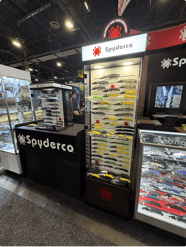 Display booth featuring Spyderco knives and various folding knives on a pegs, with additional display cases and a person in the background at an indoor trade show or exhibition.