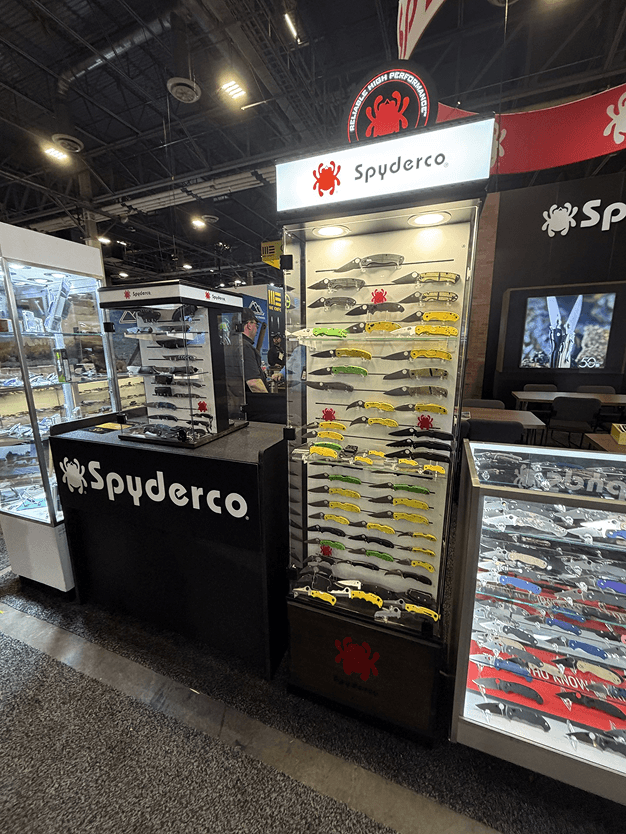 Display of model cars on a glass shelf at a Spyderco booth, with additional displays of knives and tools behind it, in an indoor convention or trade show setting.