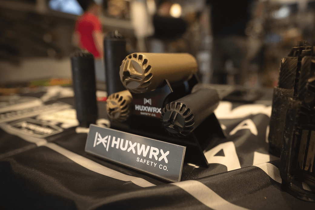 HUXWRX Safety Co. display with cylindrical, camouflage and black firearm suppressors on a table at an exhibition or trade show.