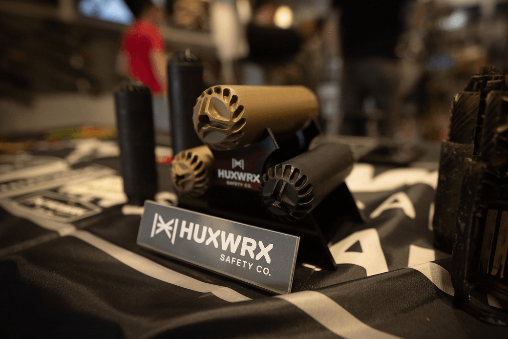 Displays firearm accessories, including silencers and packaging for HUXWRX Safety Co., on a table with a branded sign.