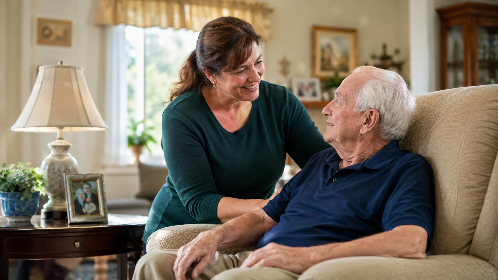 A woman smiling and holding an elderly man's hand as they sit together on a beige armchair in a cozy living room.