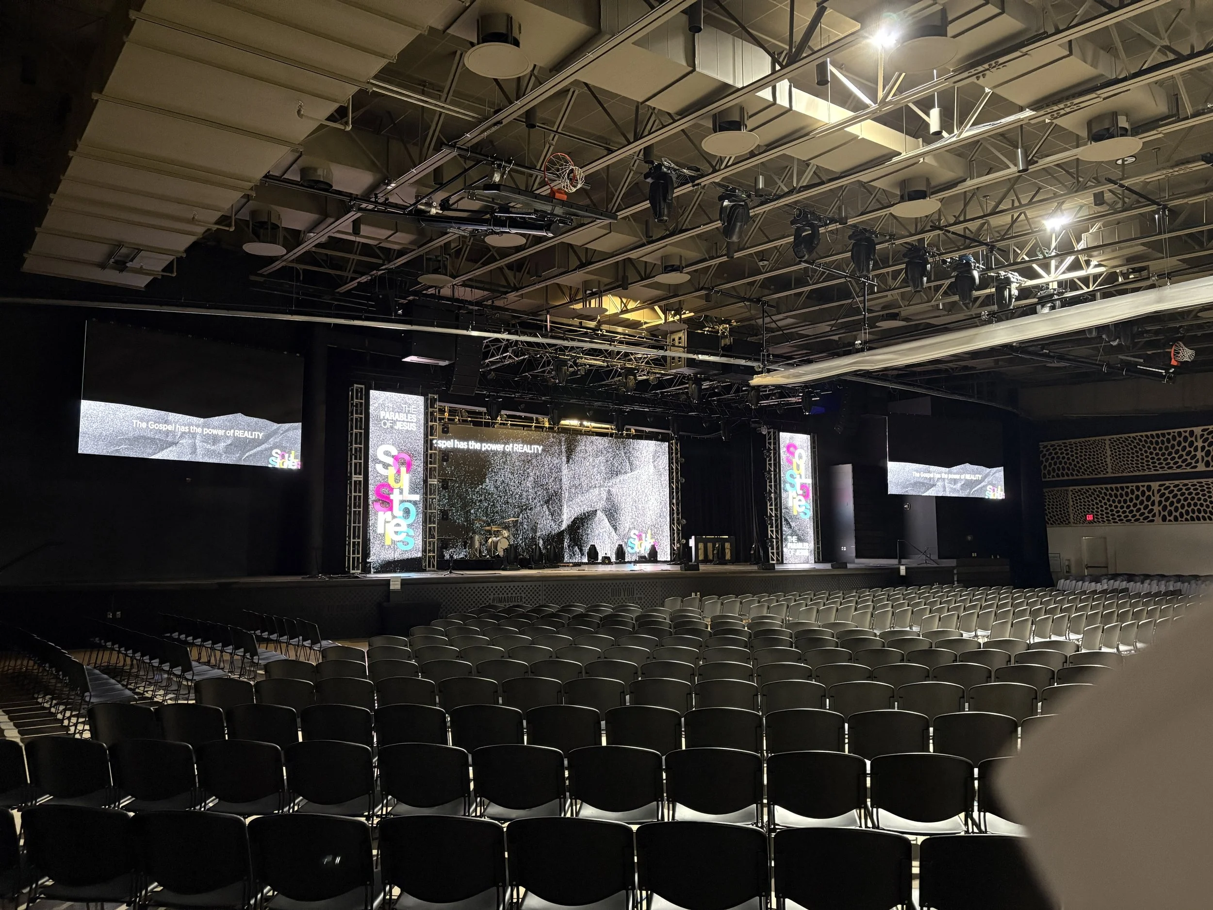 An empty auditorium or conference hall with rows of chairs facing a large stage with multiple screens displaying colorful graphics and text that reads 'The Gospel has the power of REALITY'. The ceiling has exposed beams, lighting, and equipment for sound and visuals.