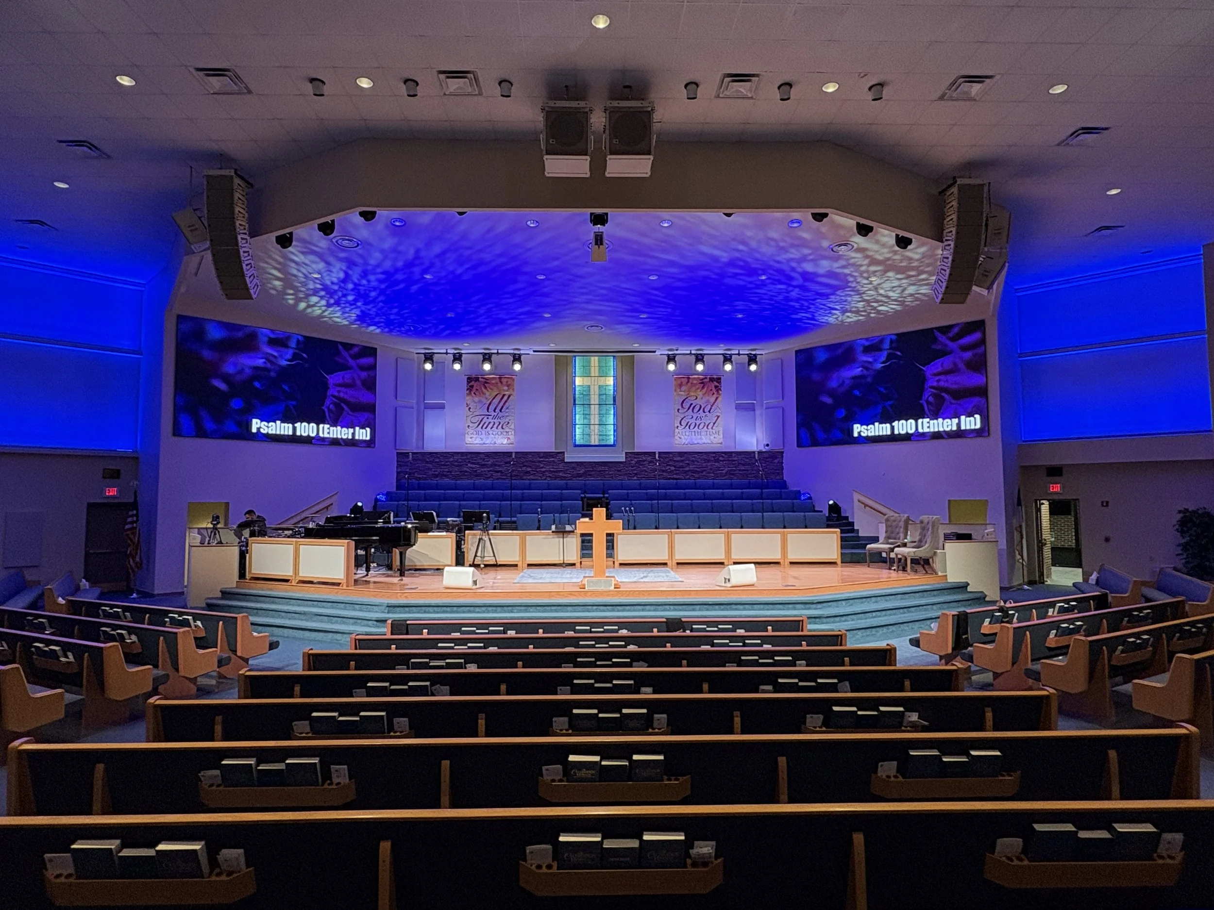 Empty church sanctuary with a stage, pulpit, grand piano, chairs, and screens displaying Psalm 100, interior lit with blue lighting.