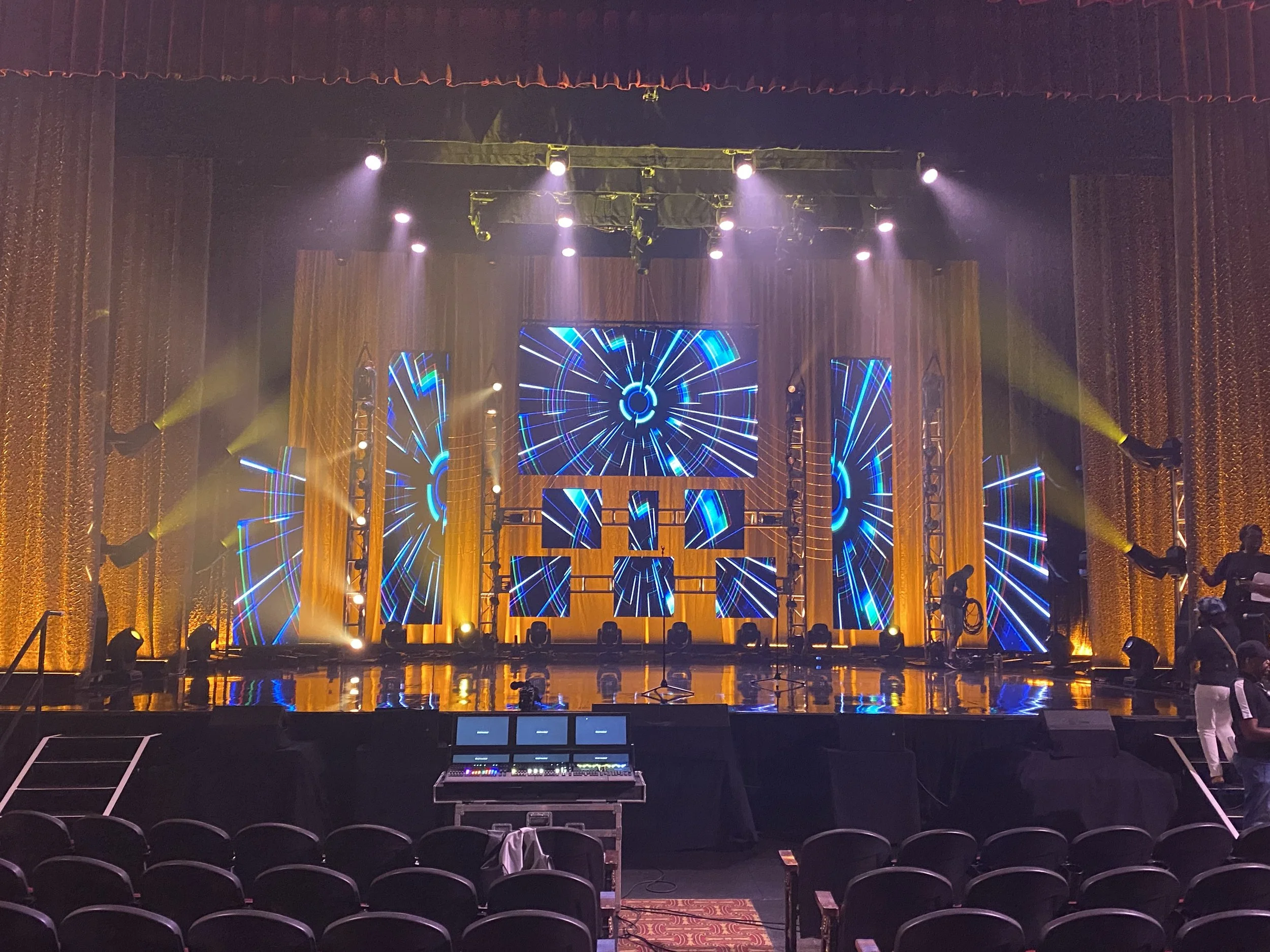Empty stage with multiple screens displaying futuristic digital designs, surrounded by yellow and purple lighting, and staged for a concert or event.