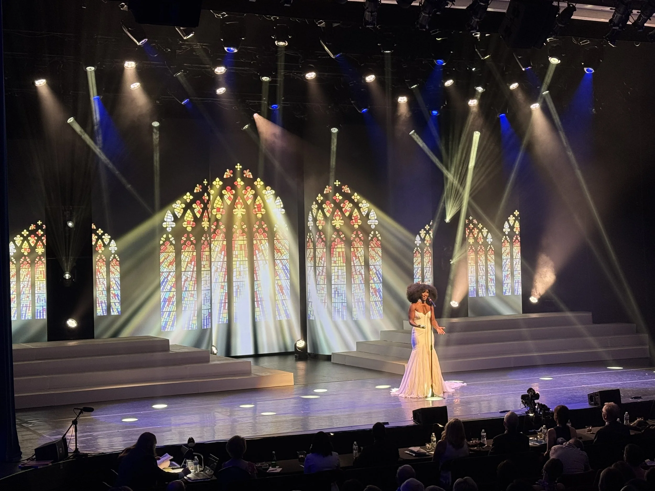 A woman in a long, elegant gown performs on stage with stained glass window backdrops and stage lighting, while an audience watches from below.