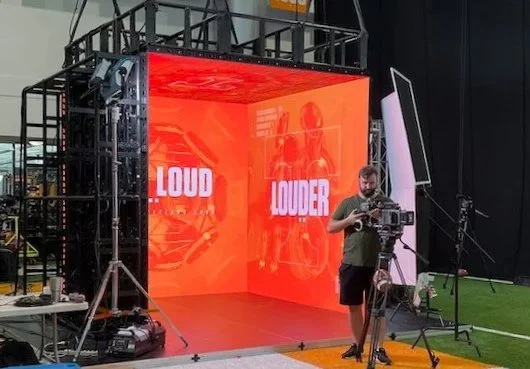 A man operating a video camera on a green field next to a large LED screen displaying the words 'LOUD' and 'LOUDER' in red and white.