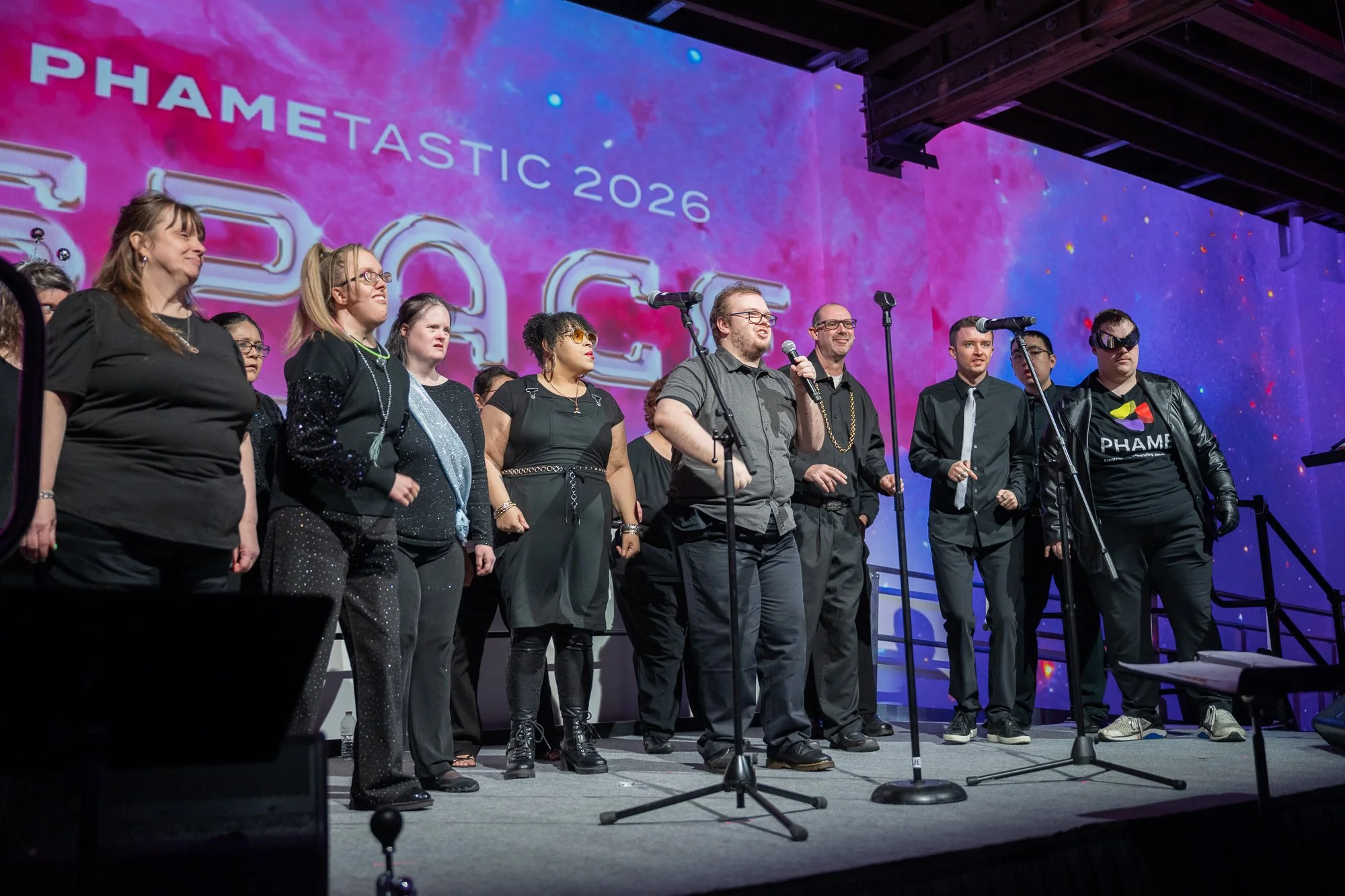 A group of performers dressed mostly in black stand on stage with microphones in front of a colorful “PHAMEtastic 2026” backdrop.
