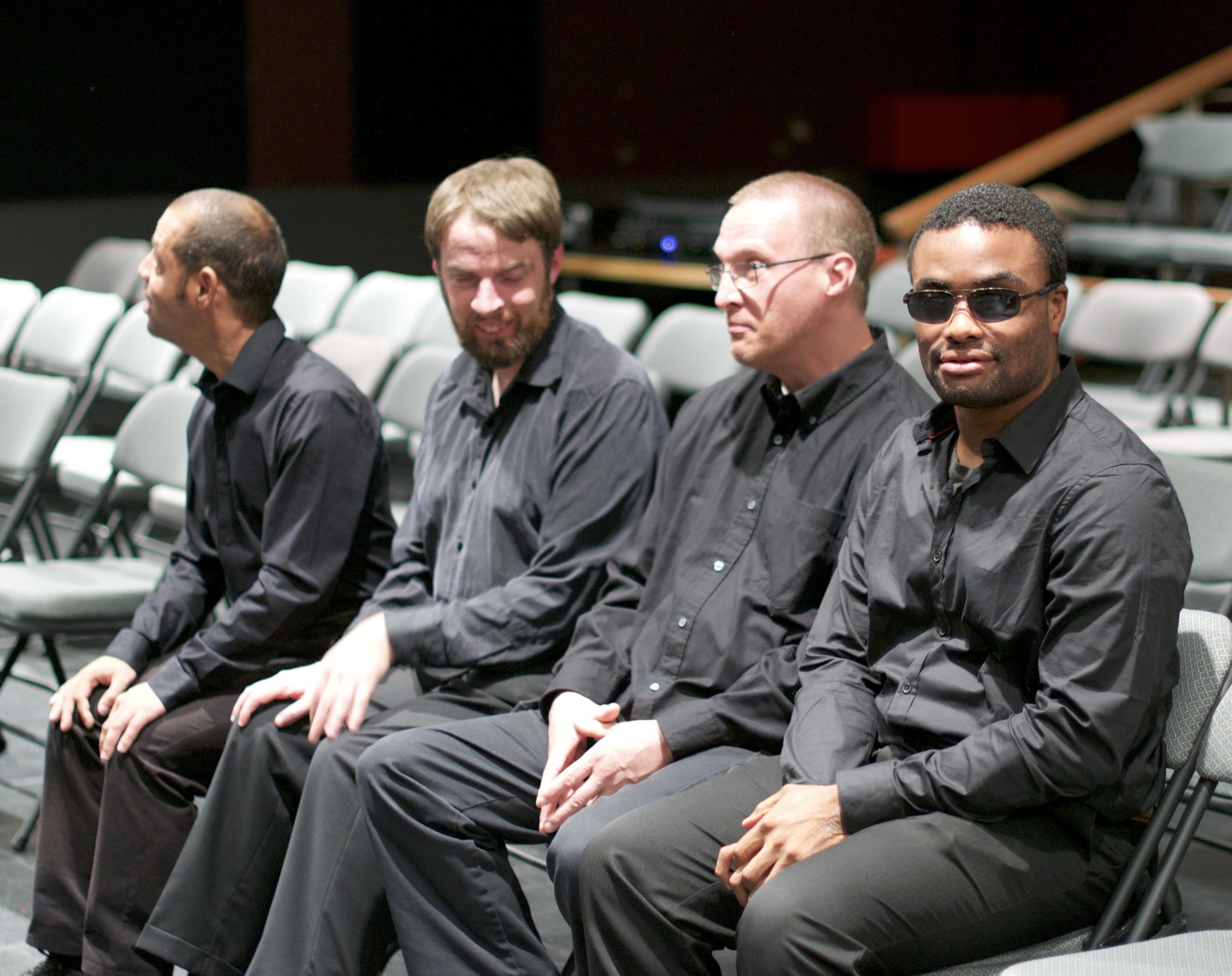 Four men sitting in folding chairs, wearing all black