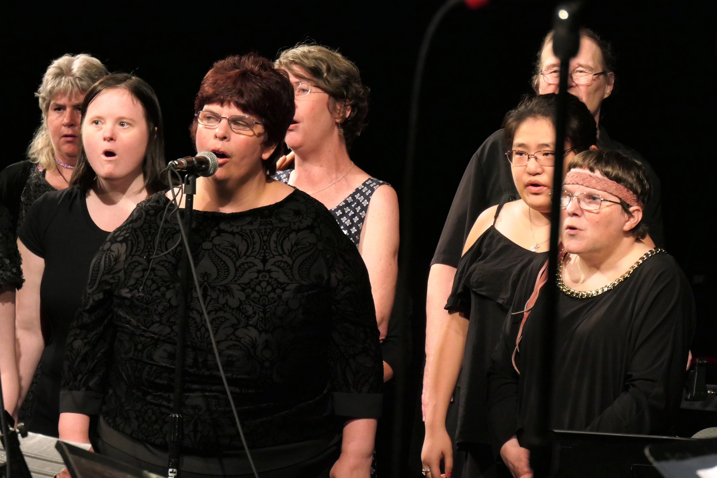 Group of women singing , some with microphones, in a dark room