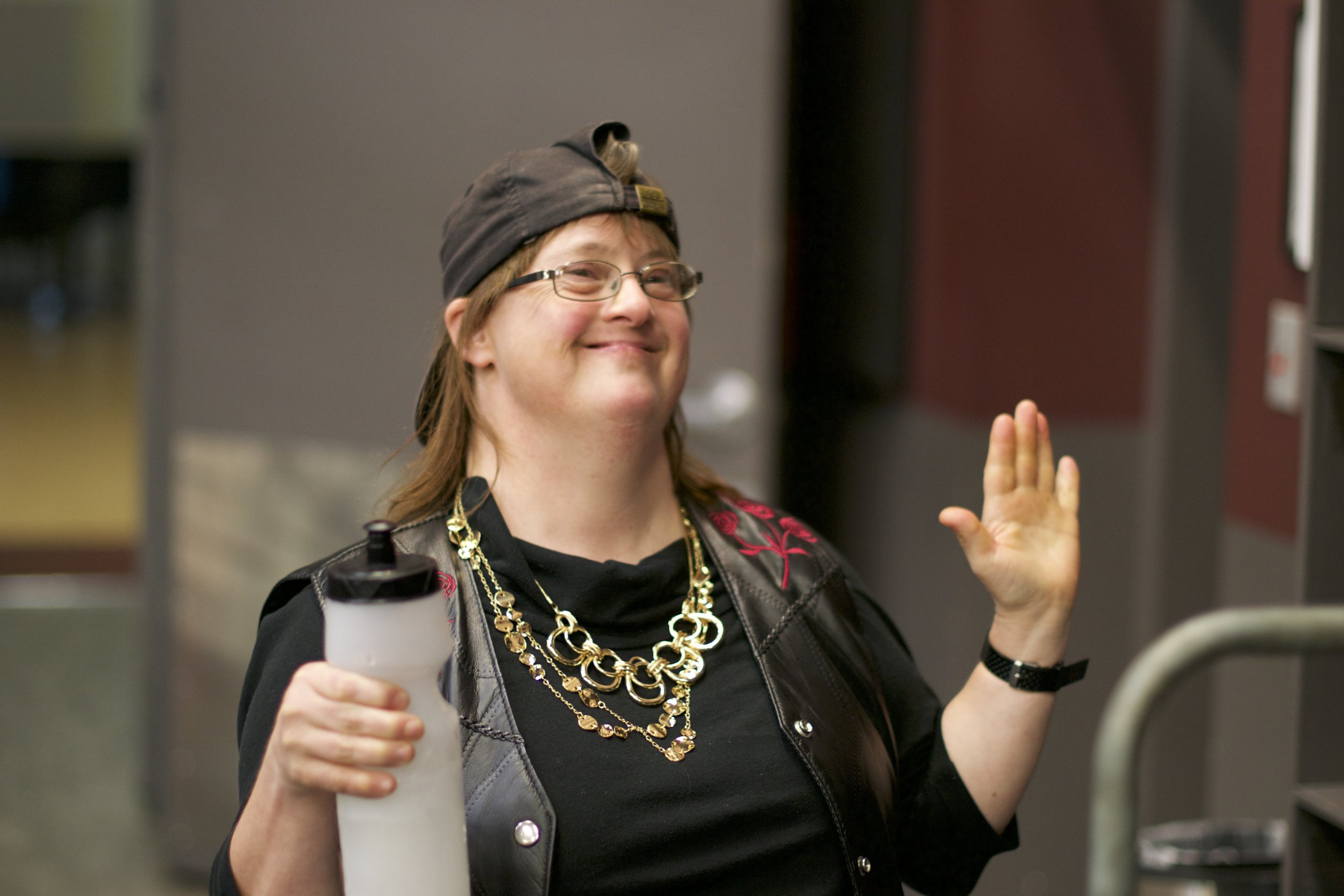 Smiling woman wearing backwards cap and gold necklace, holds a coffee cup and raises the other hand