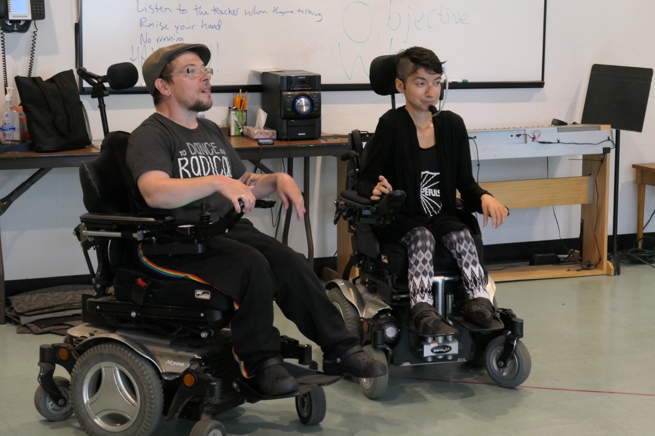 Two people wearing dark fashionable clothes seated in wheelchairs.