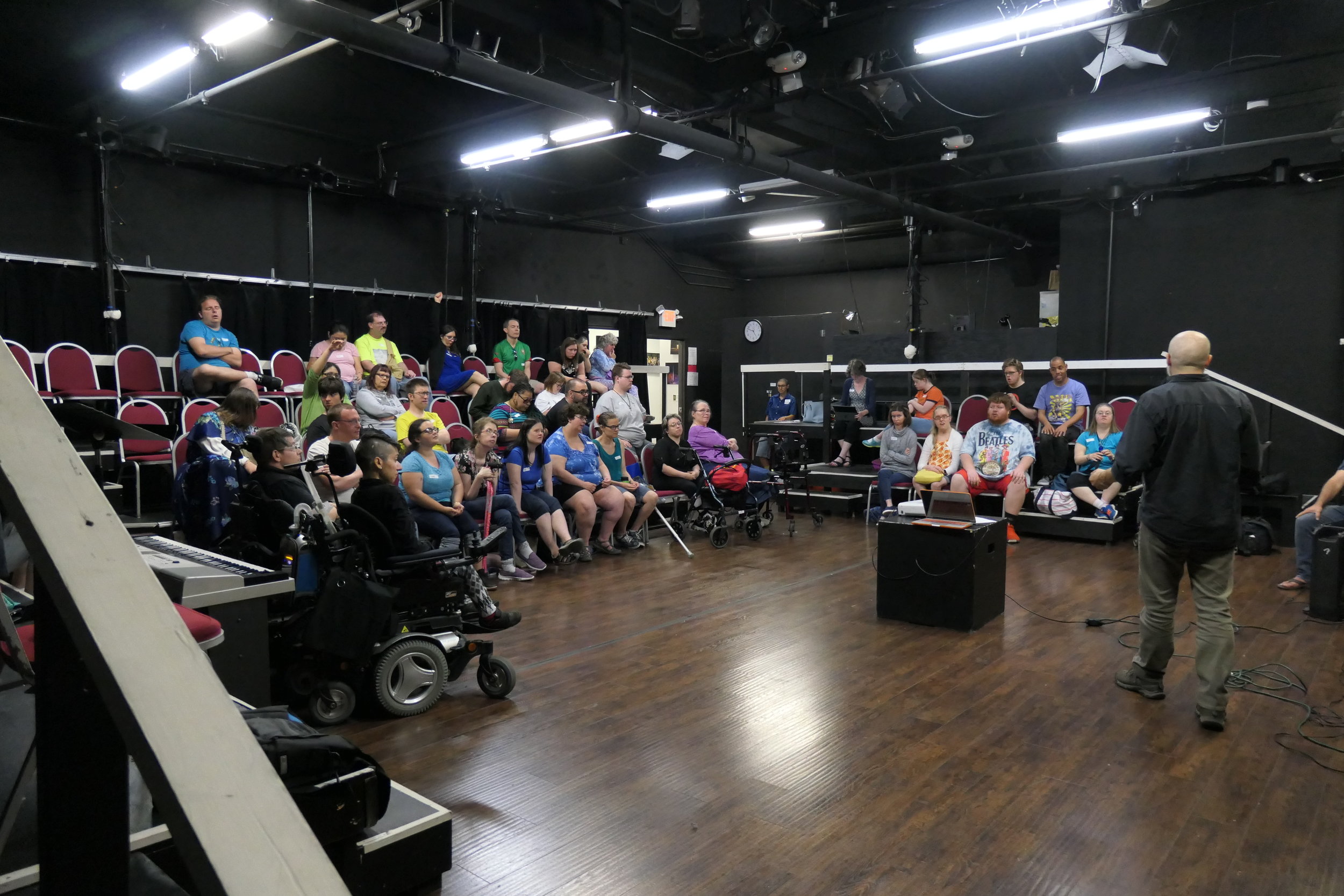 Man speaking to a large group of seated people in a black box theatre