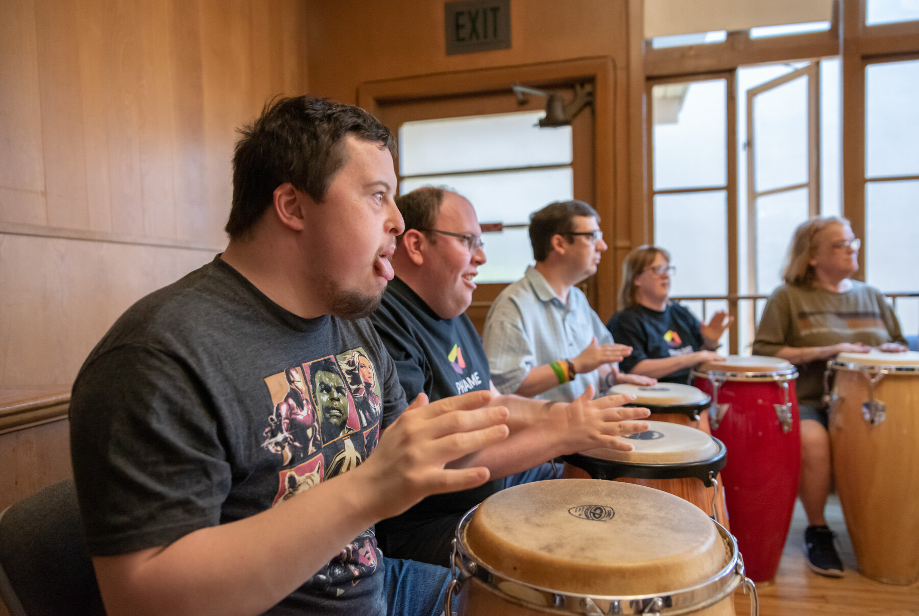 Line of people playing drums, the young man in front has his tongue out in concentration.