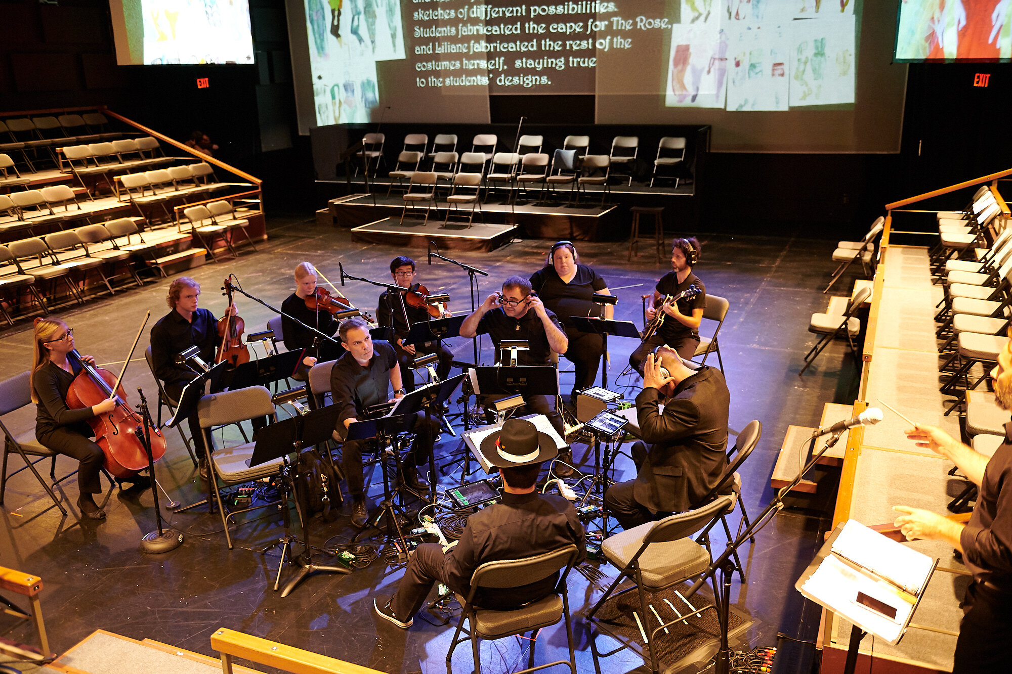 Group of people seated on the stage of a black box theatre playing instruments.