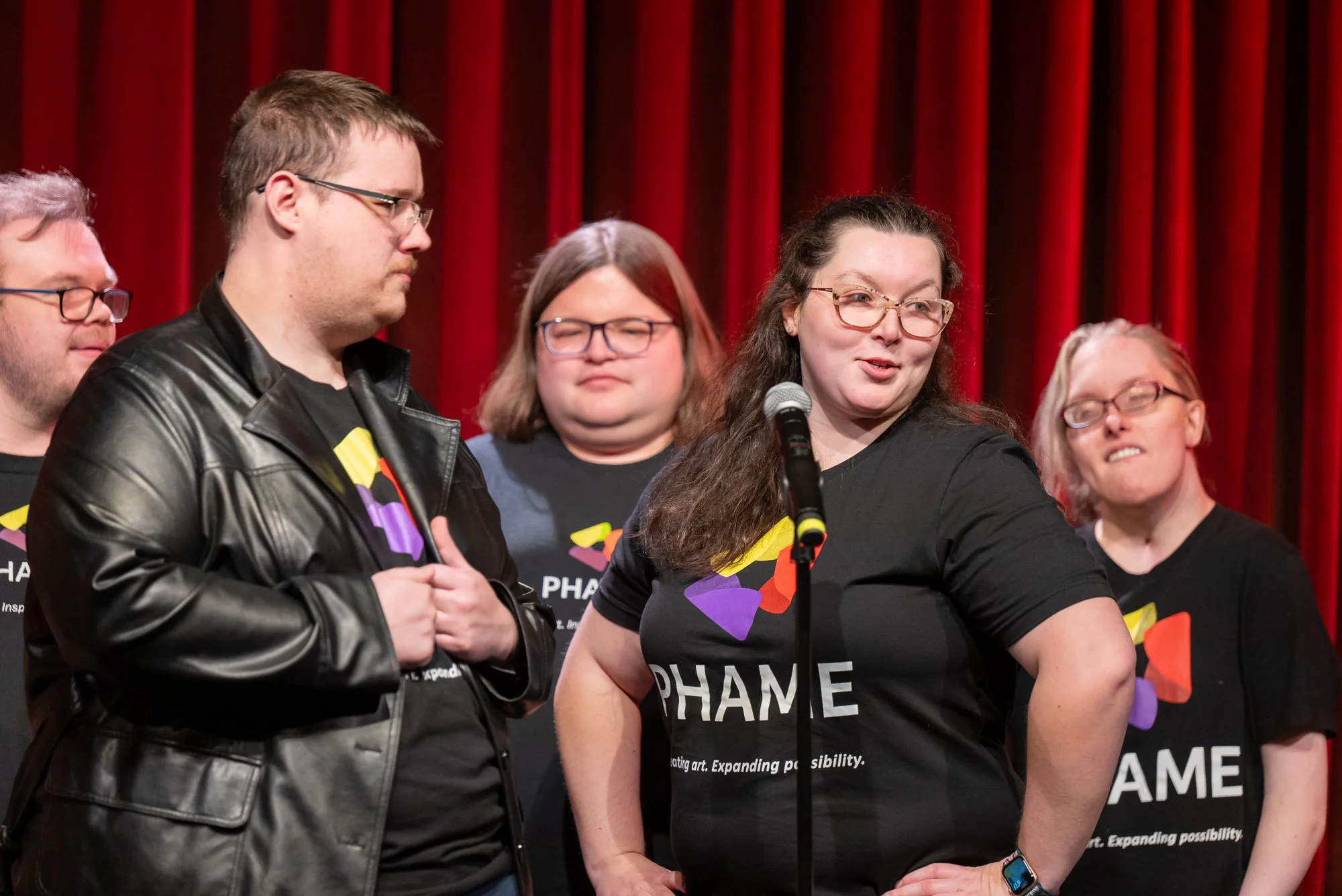 Five people gathers in front of a mic and a dramatically lit red curtain.