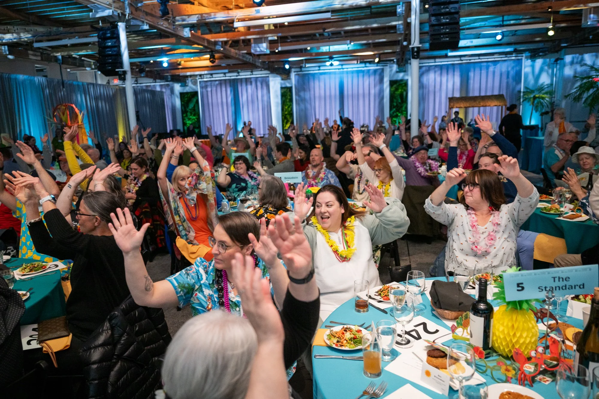 Large crowd of seated people raising their arms like they're doing the wave. They wear tropical attire.