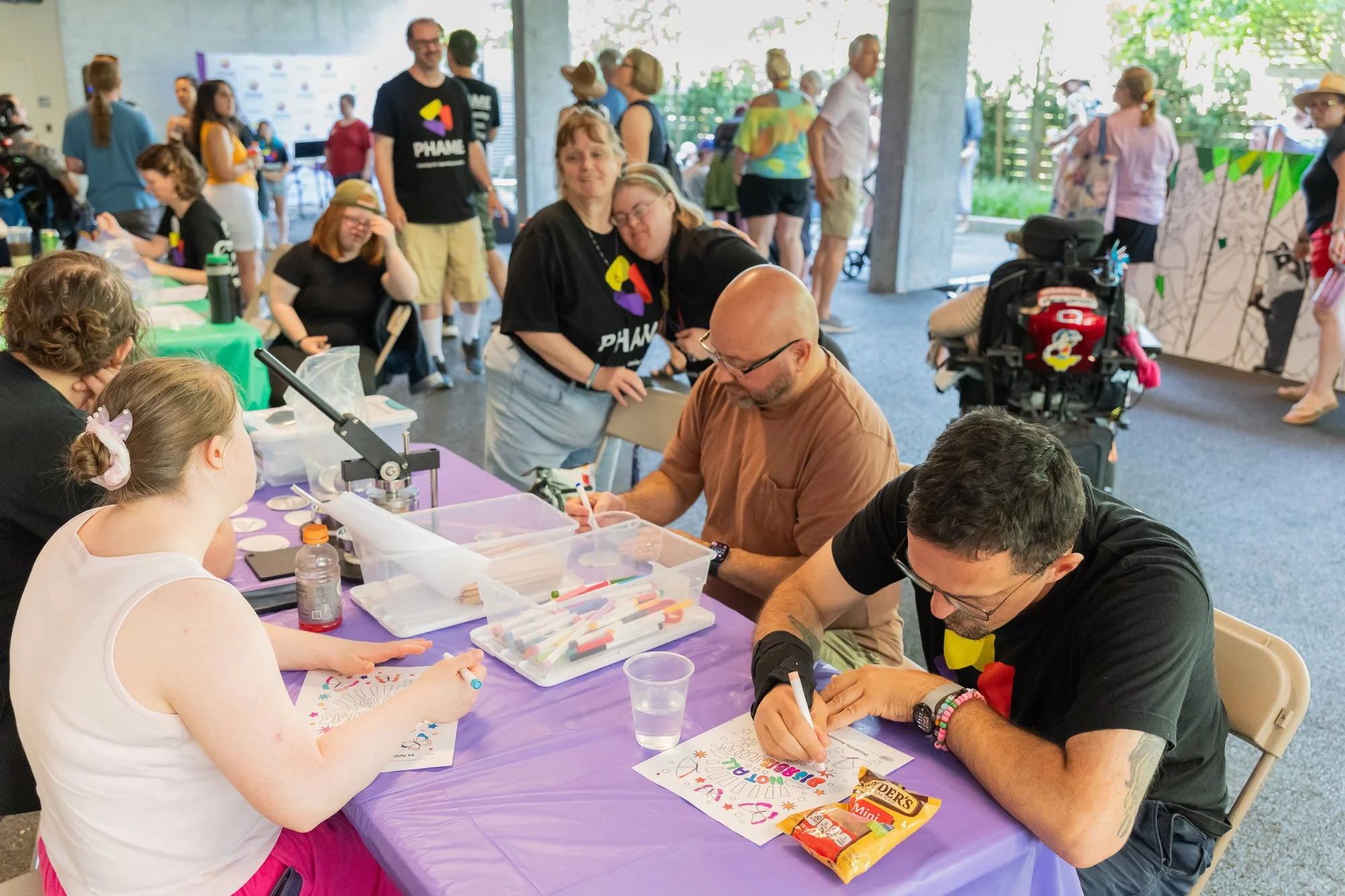 Multiple people sitting at a crafts table and working on art pieces. Behind them is a crowd of people mingling.