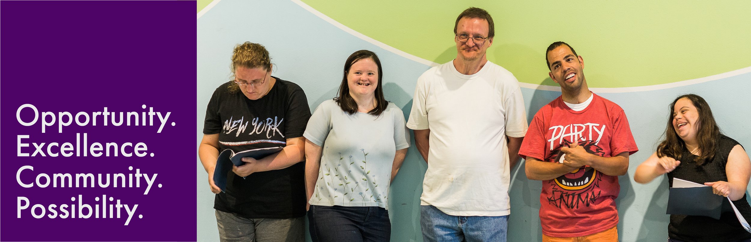 Five people standing against a wall painted with blue, green, and white. One person looks down at an open folder, another holds an open folder and gestures playfully. On the left are the words Opportunity, Excellence, Community, Possibility.