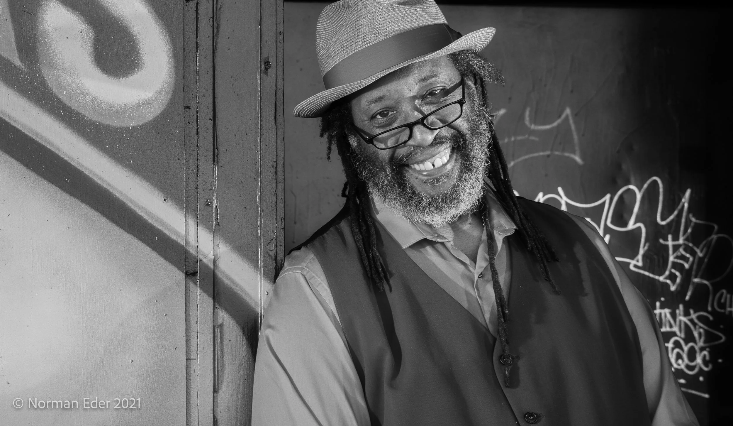Smiling man with a beard and long dreads wearing glasses, a hat, and a vest over a collared shirt.