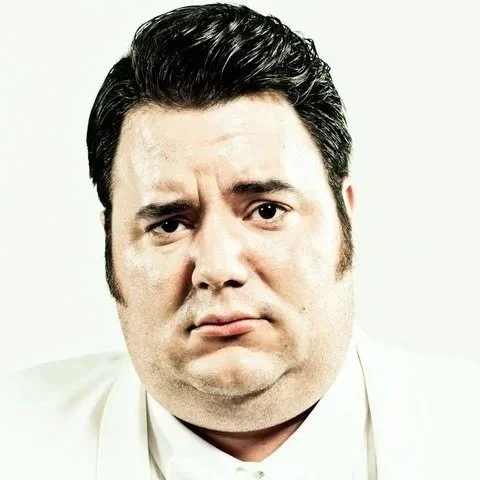 Man with slicked back dark hair and a furrowed brow wears a white suit that blends with the white background.