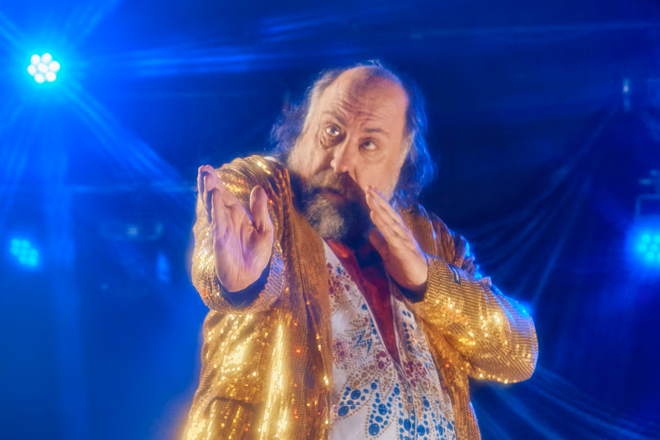 A man with a beard doing a karate chop pose while performing in a gold sequin jacket in front of a blue backlit stage.
