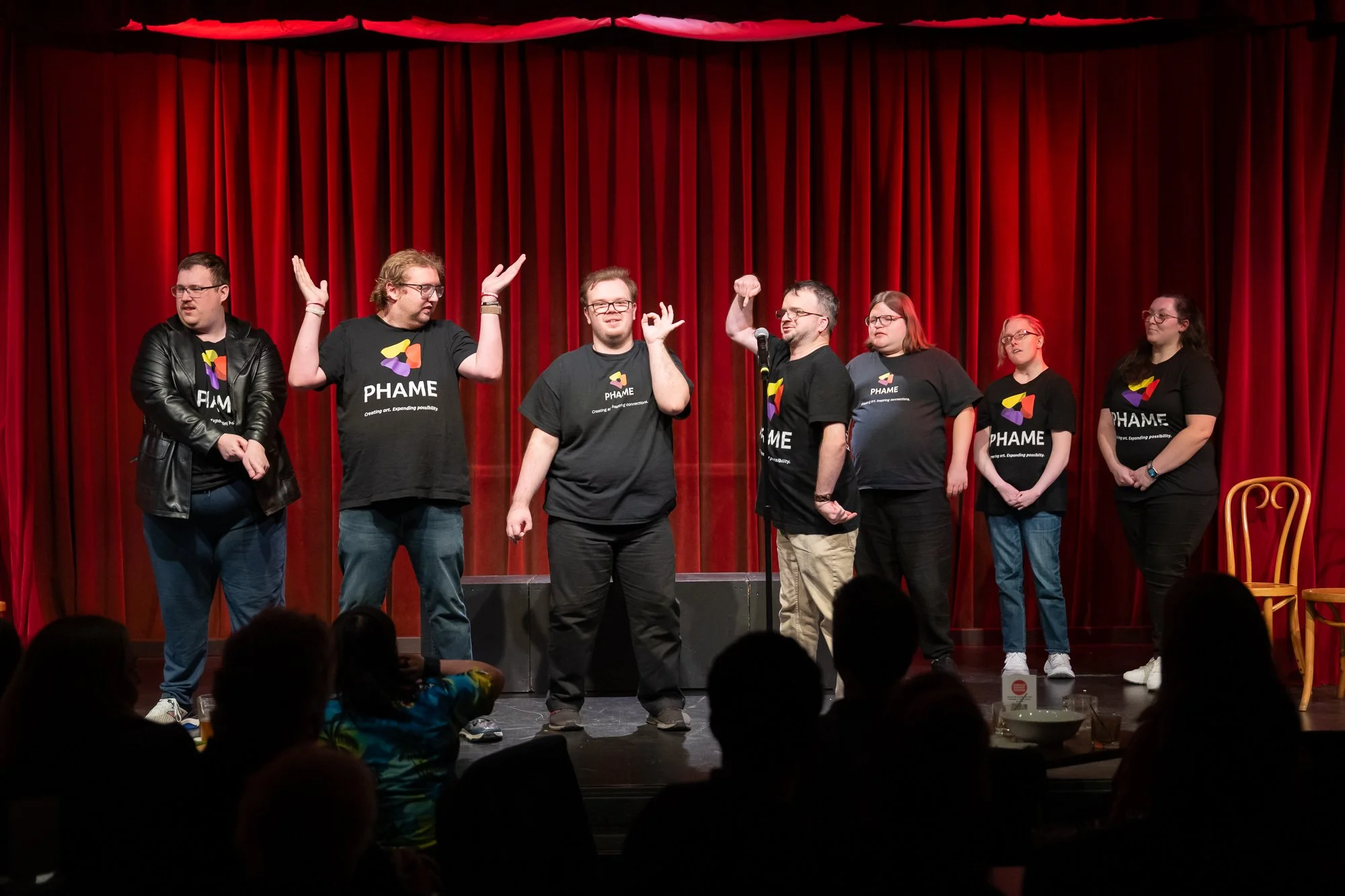 Seven performers wearing PHAME shirts on a stage. Behind them is a red curtain. Three of them pose comedically.