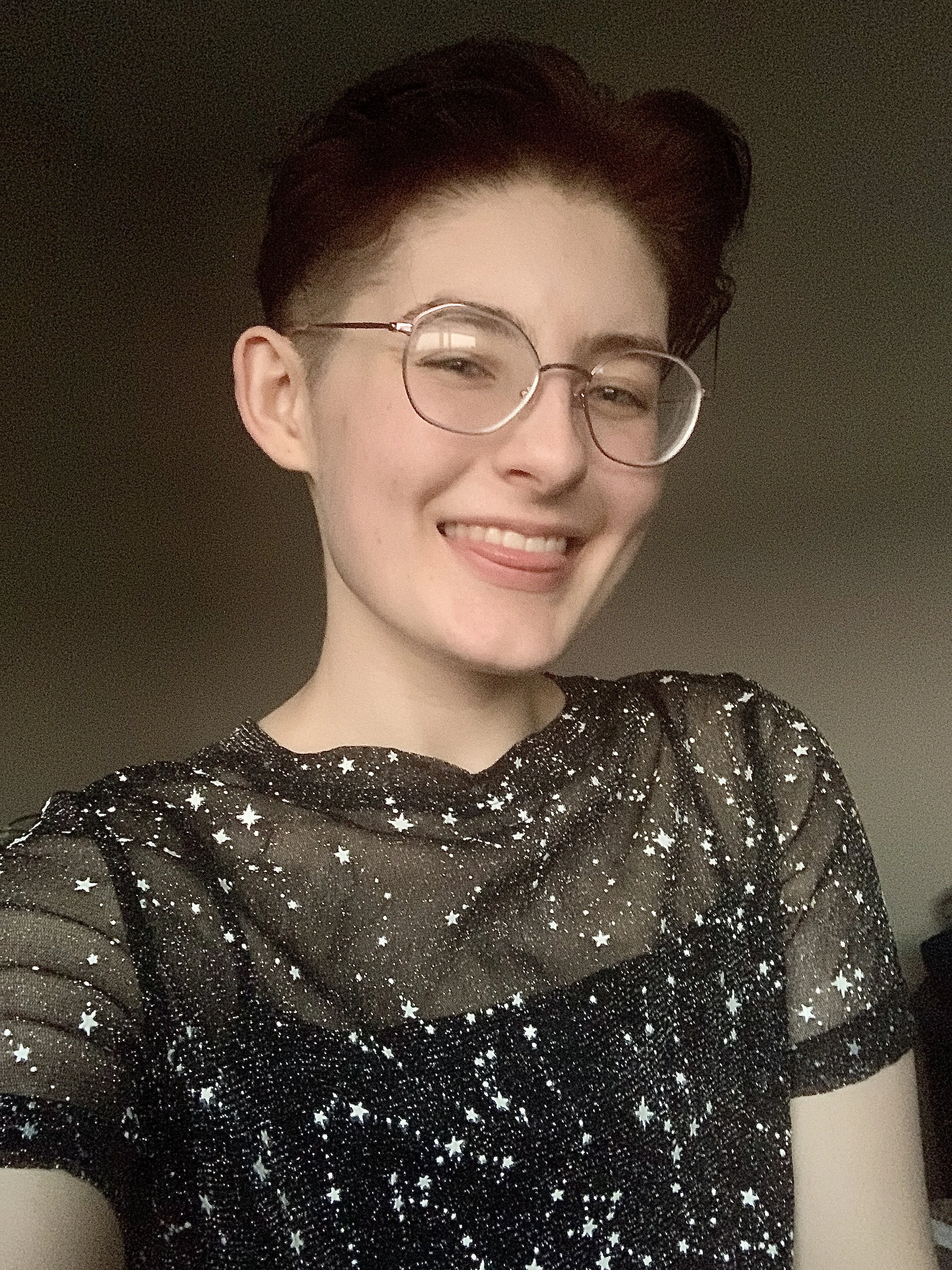 Person with short dark hair and glasses wearing a dark shirt with light colored stars on it. They smile and show their tongue a little.