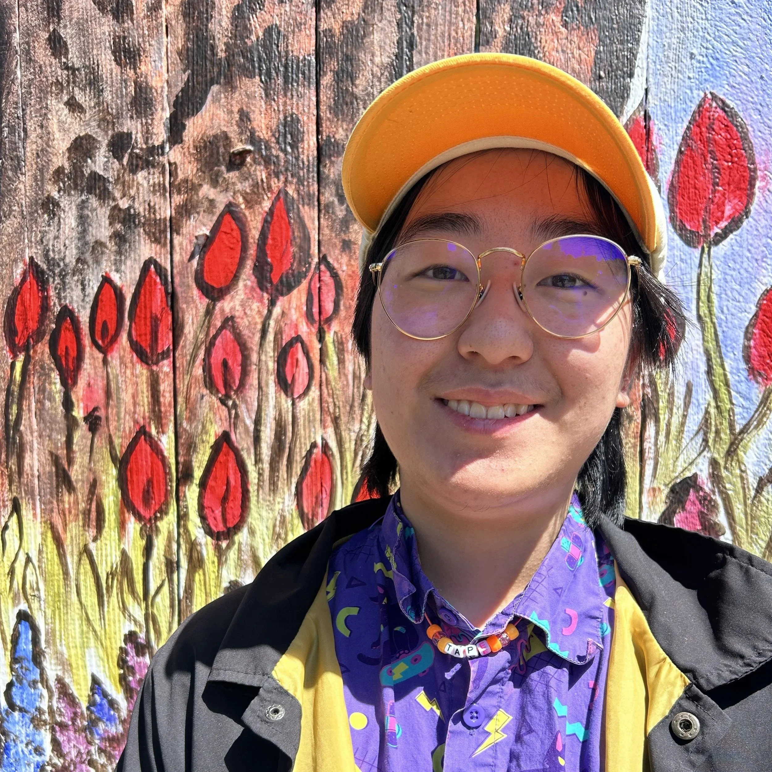 Photo of a smiling person with a yellow hat, glasses and a purple shirt in front of a colorful floral background.