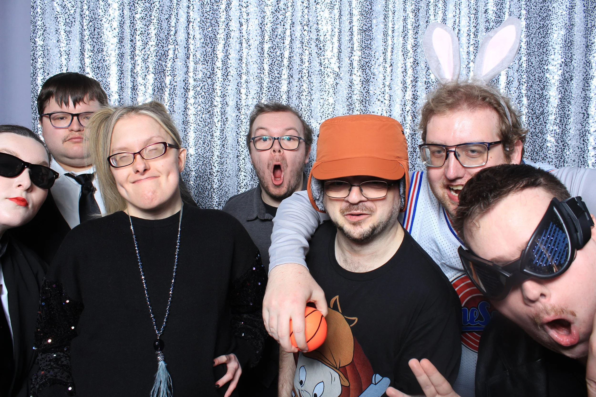Seven people in dark clothing pose closely together in front of a silver sequin backdrop, some wearing glasses and novelty props.