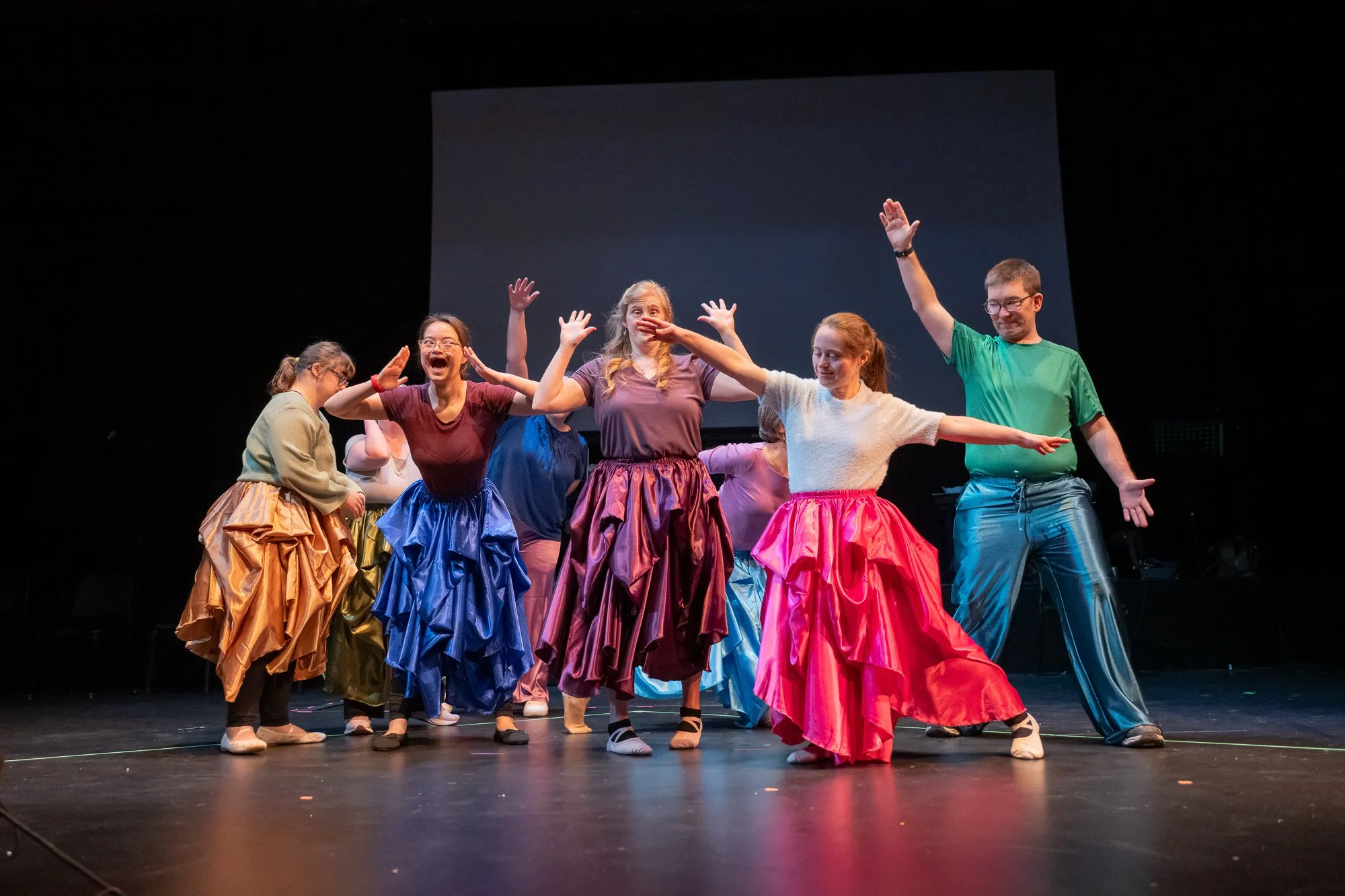 Eight dancers on a dramatically lit black stage. They wear shiny colorful outfits and pose theatrically.