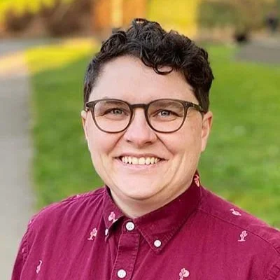 Person with short dark curly hair wears glasses and a crimson collared shirt. Behind them is grass and a sidewalk out of focus.