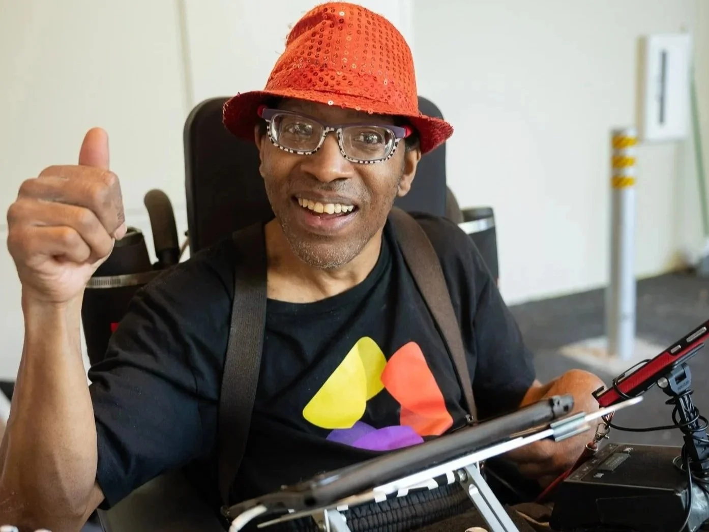 A person wearing a red sequined hat and glasses gives a thumbs-up while seated in an adaptive chair with mounted equipment and a tablet device.