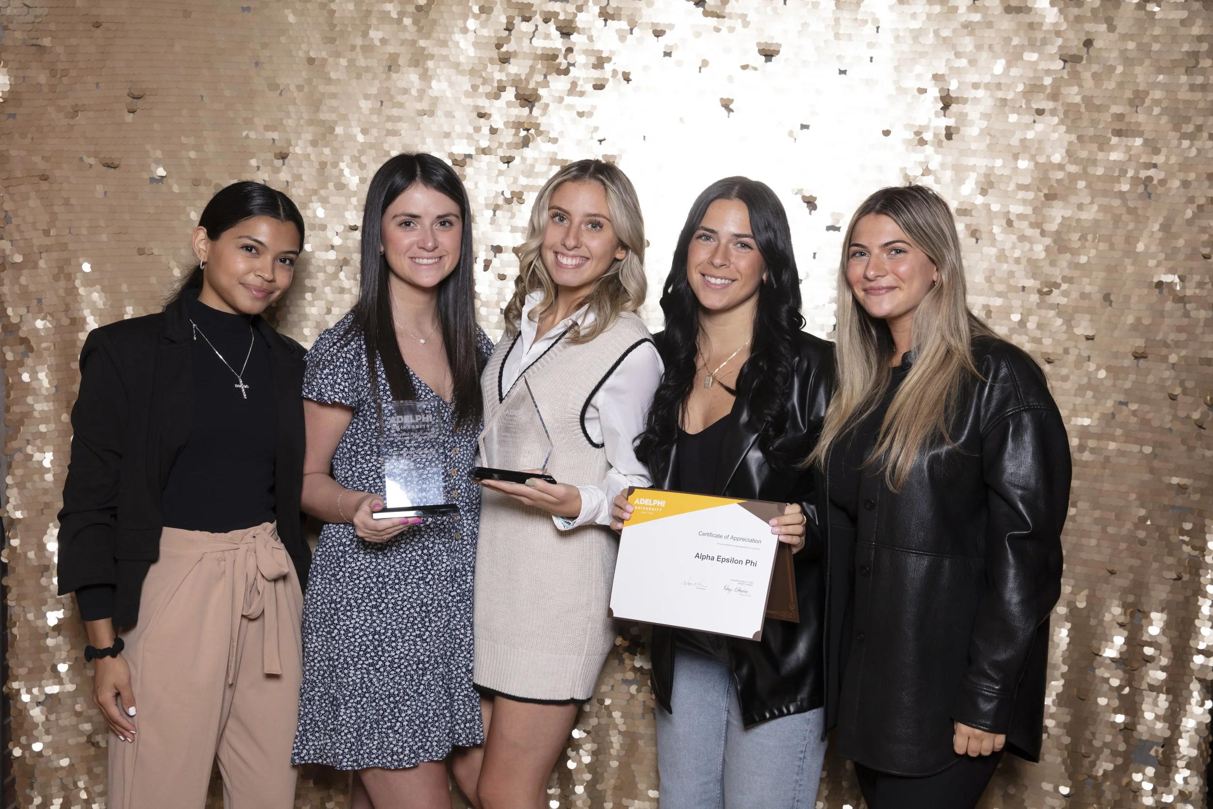 Group of five young women standing in front of a gold sequin backdrop, holding awards and a certificate, smiling for the camera at an event.