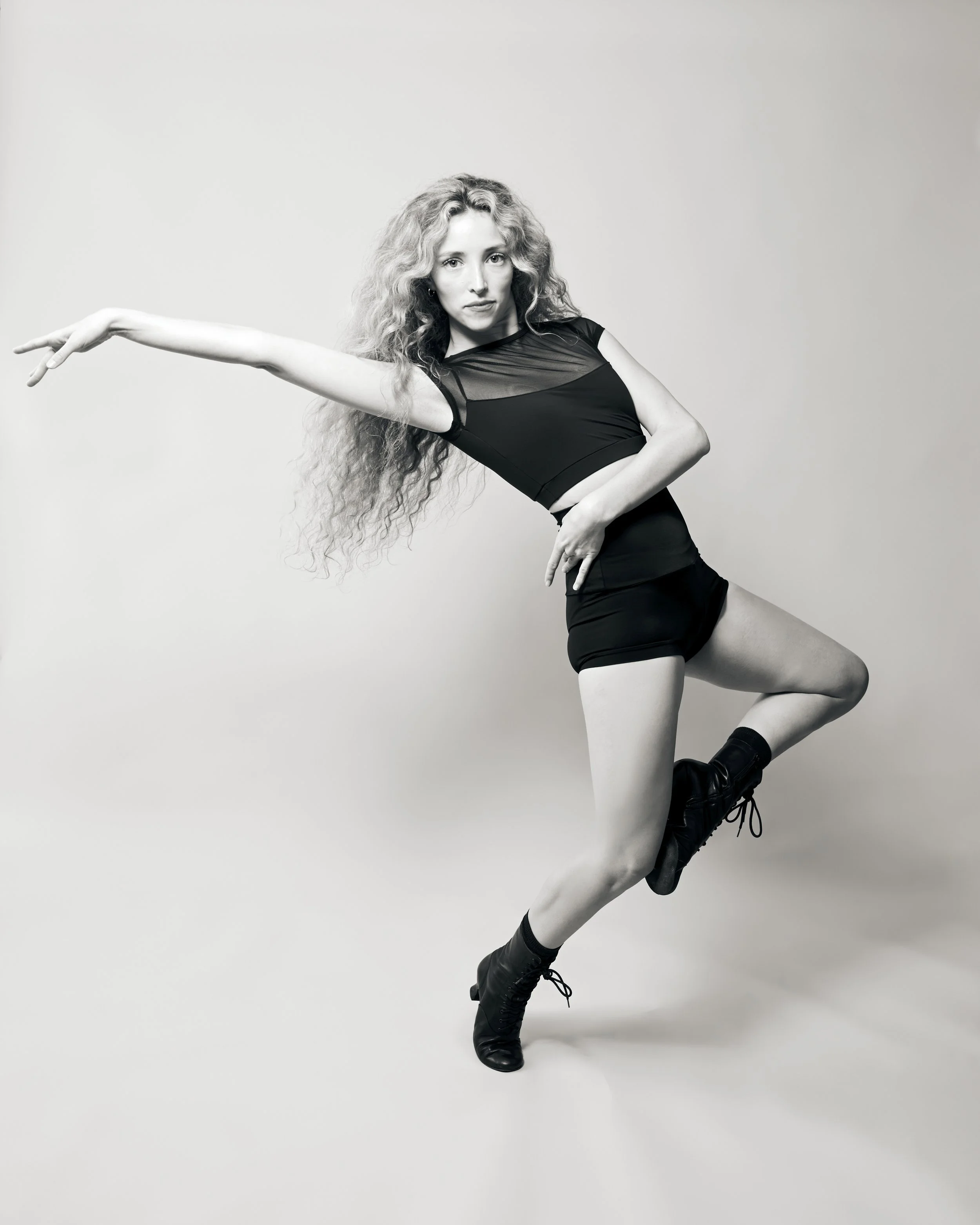 Black and white photo of a woman with long curly hair, wearing a fitted black top with sheer details, a black skirt, and lace-up black boots. She is dancing or posing dynamically against a plain light background.