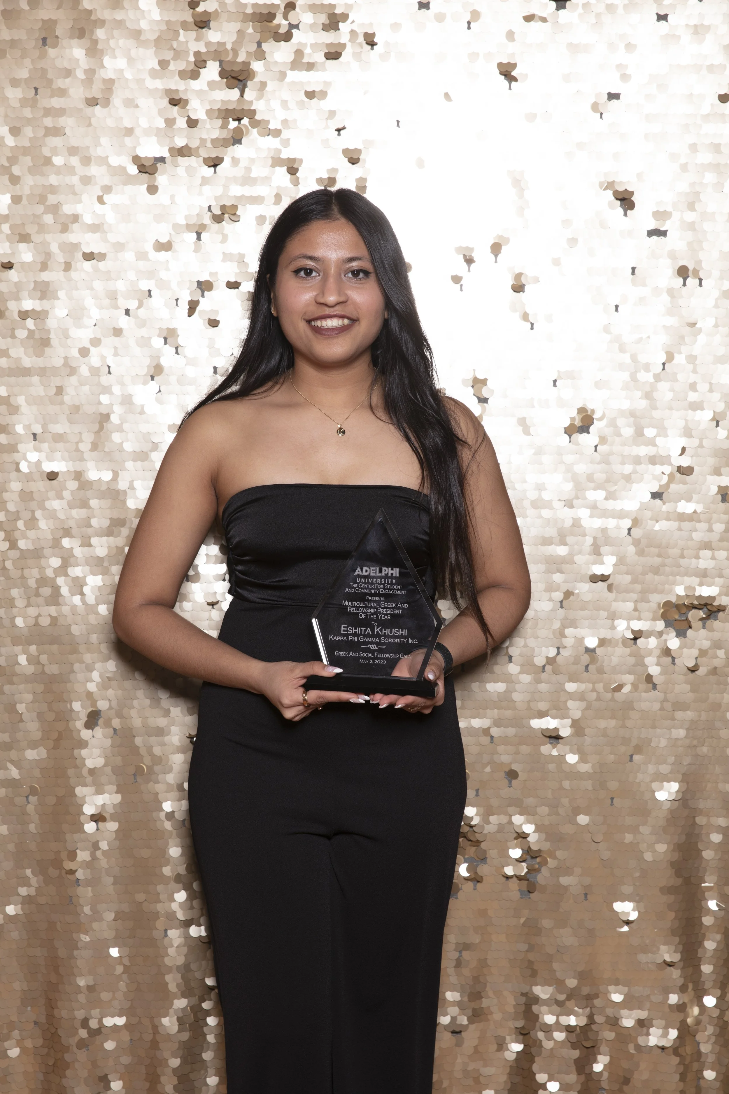 Young woman in black dress holding an award, standing against a gold sequin backdrop at an event.
