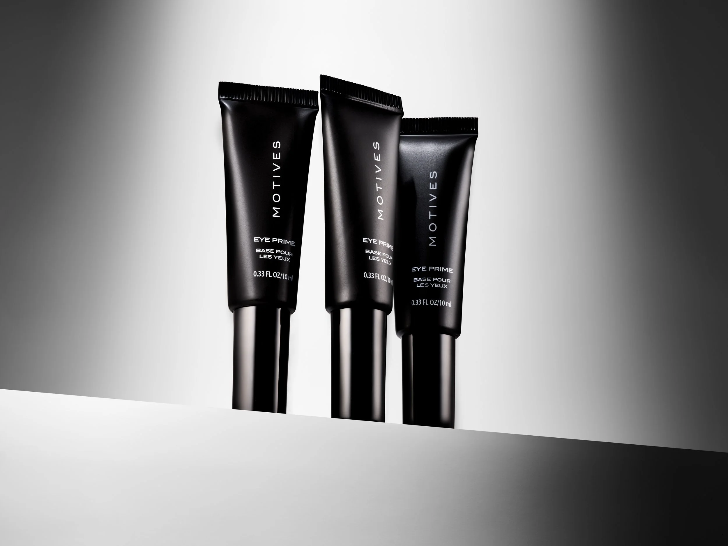 Three black tubes of MOTIVES Eye Prime Base for Eyes on a white and gray background.
