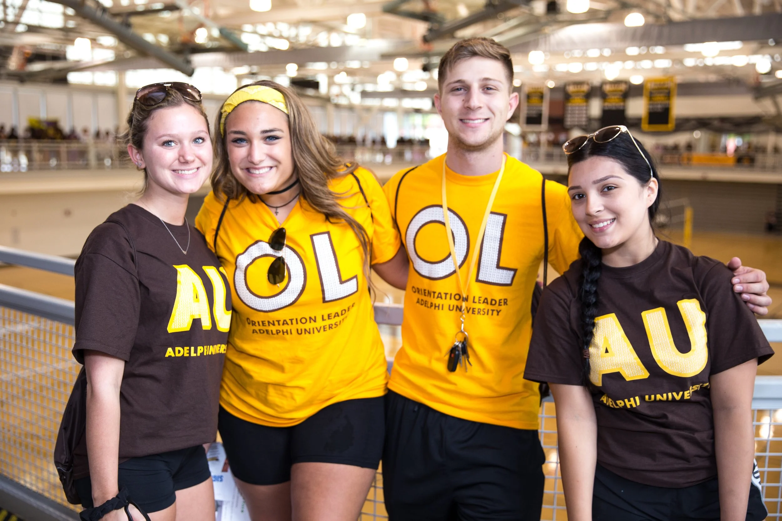 Group of four young people smiling, two wearing yellow shirts and two wearing brown shirts, in an indoor sports or event venue.
