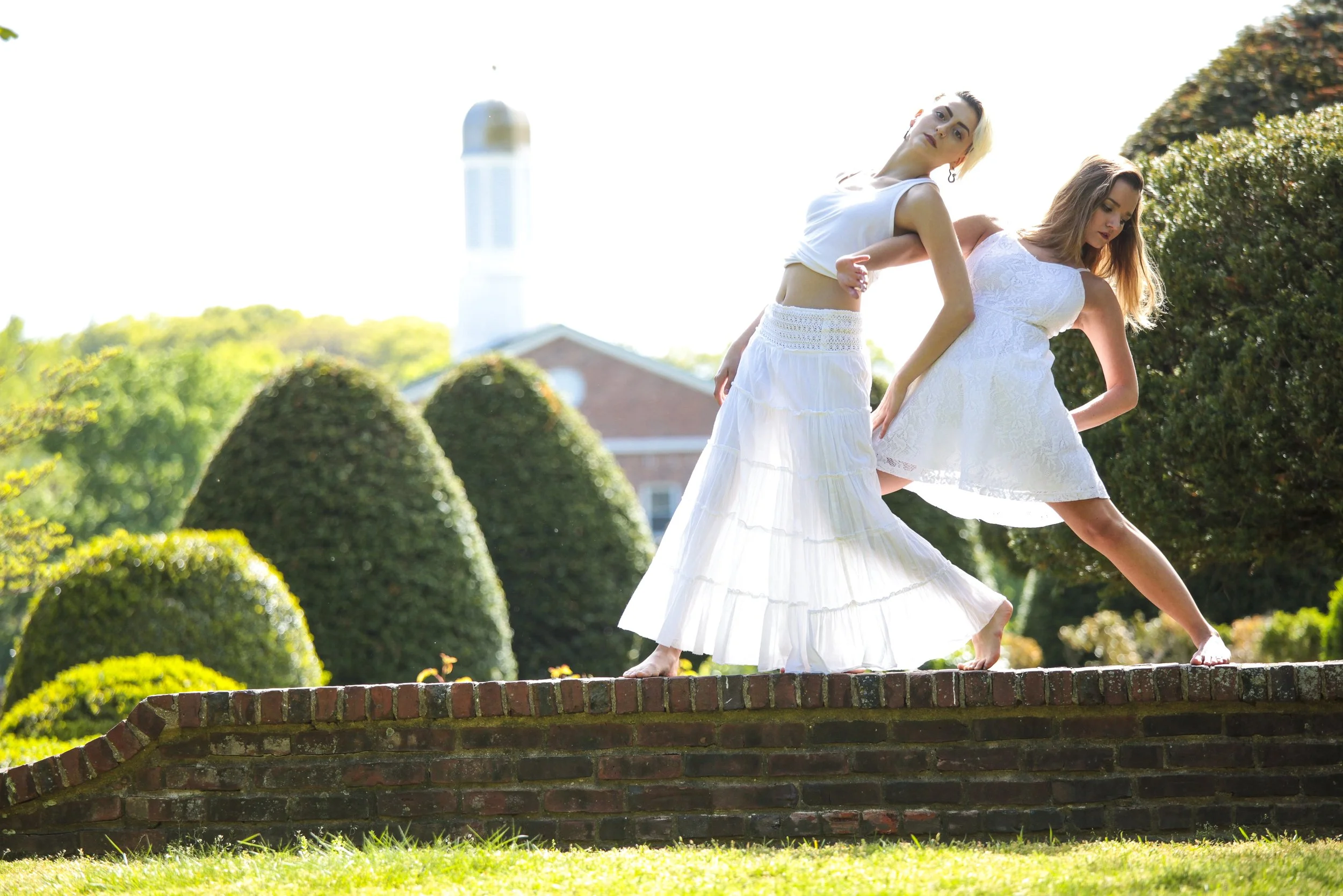 Two women in white dresses standing on a brick wall outdoors with manicured bushes and a white building with a tower in the background.
