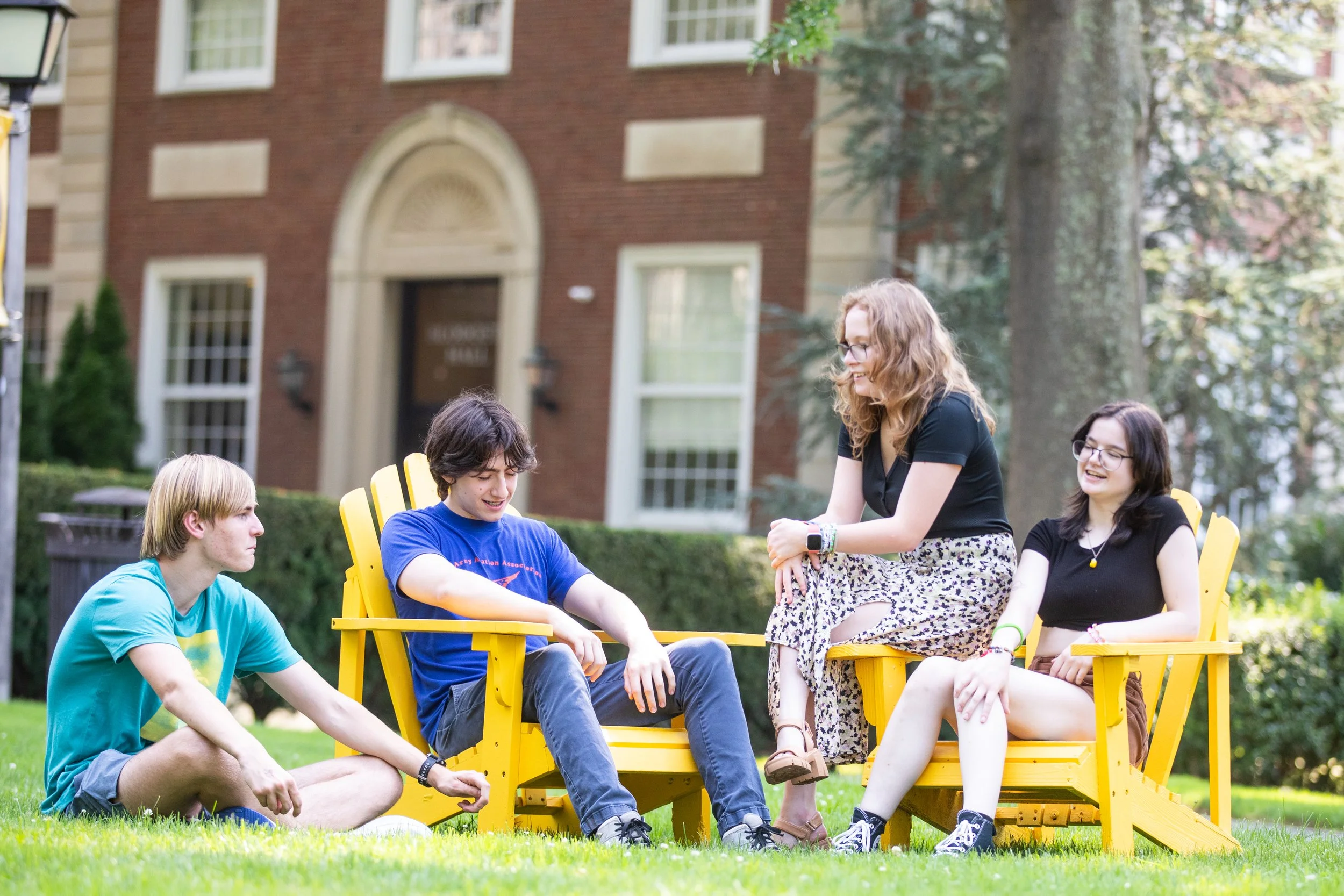 Four young people sitting outdoors on yellow wooden chairs and a blanket, engaging in conversation with smiles.