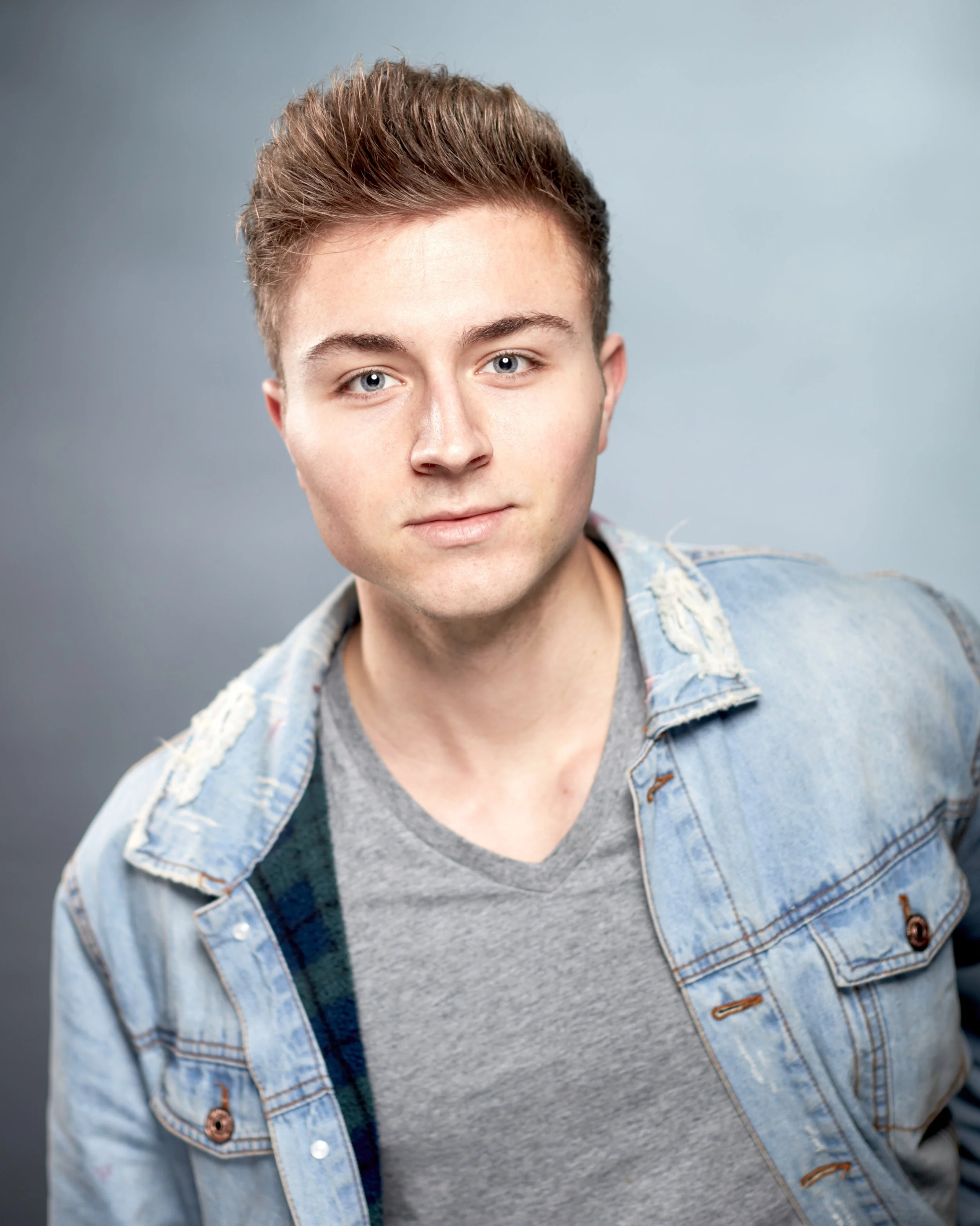 Portrait of a young man with short light brown hair, blue eyes, wearing a light blue denim jacket with a grey t-shirt underneath, against a plain grey background.