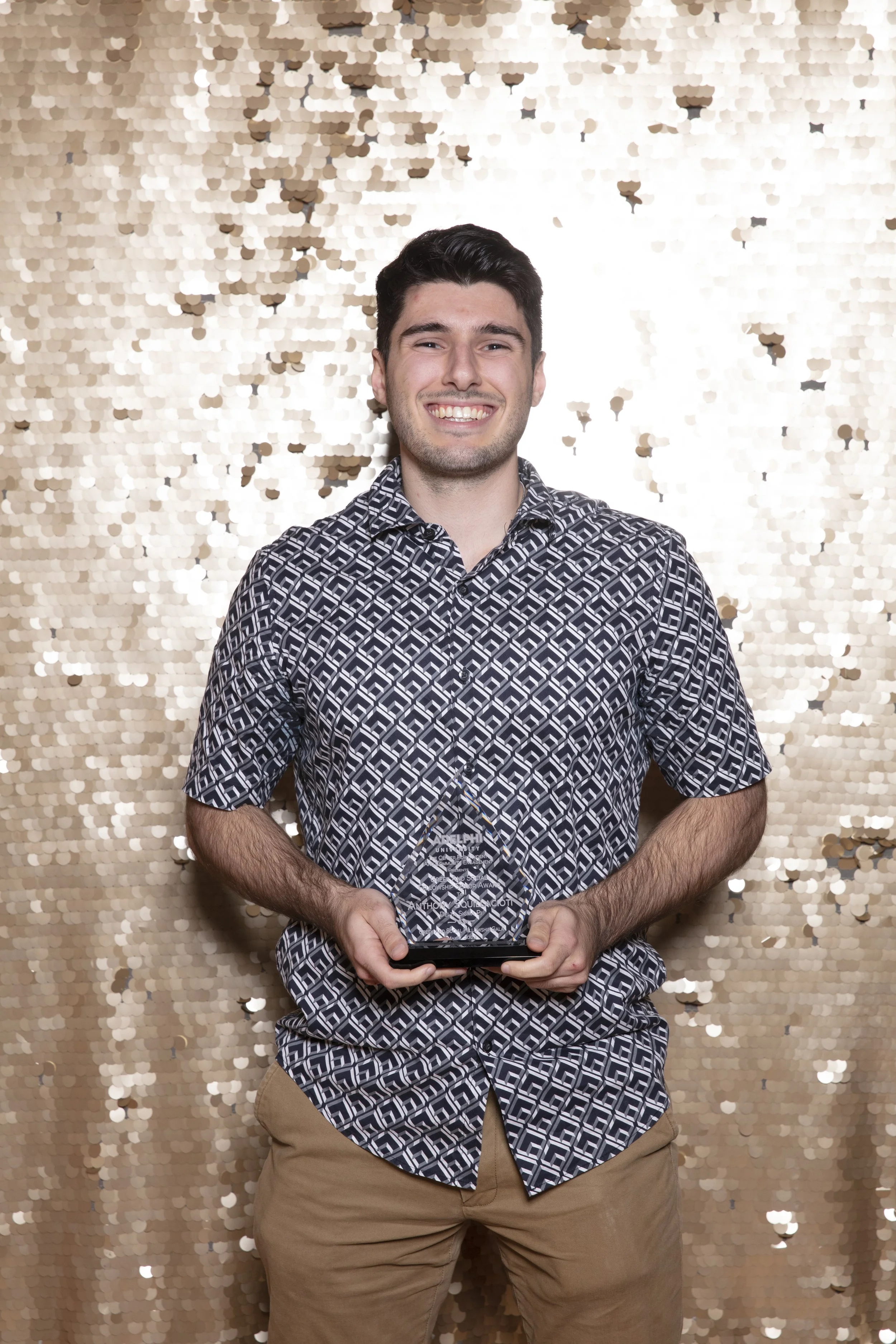 A young man smiling, holding an award or trophy, standing in front of a gold sequin backdrop.