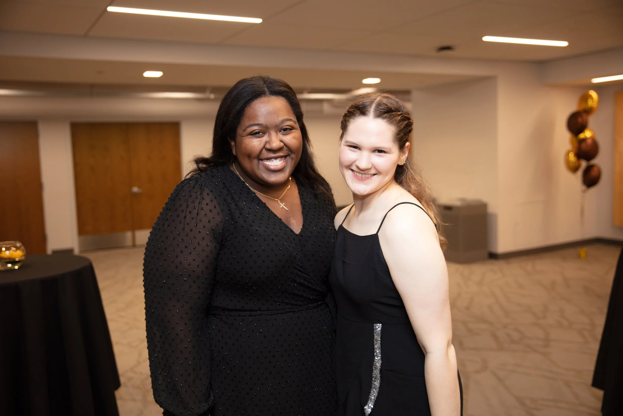 Two smiling women standing together at an indoor event, both wearing black dresses, with one wearing a black dress with sheer sleeves and the other a sleeveless dress with a silver stripe detail.