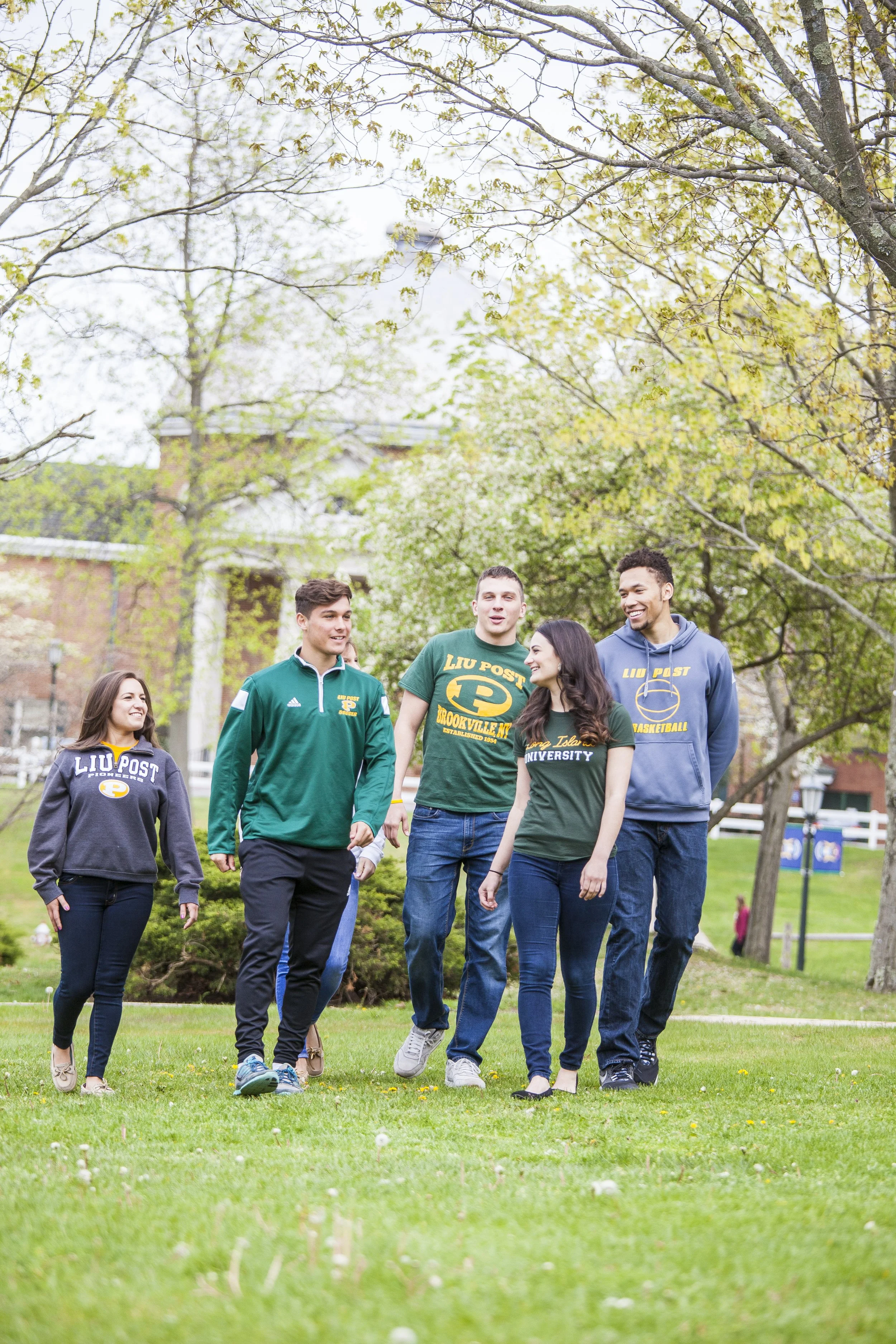 Group of five college students walking and talking outdoors on a grassy area with trees in the background, wearing University of Pittsburgh apparel.