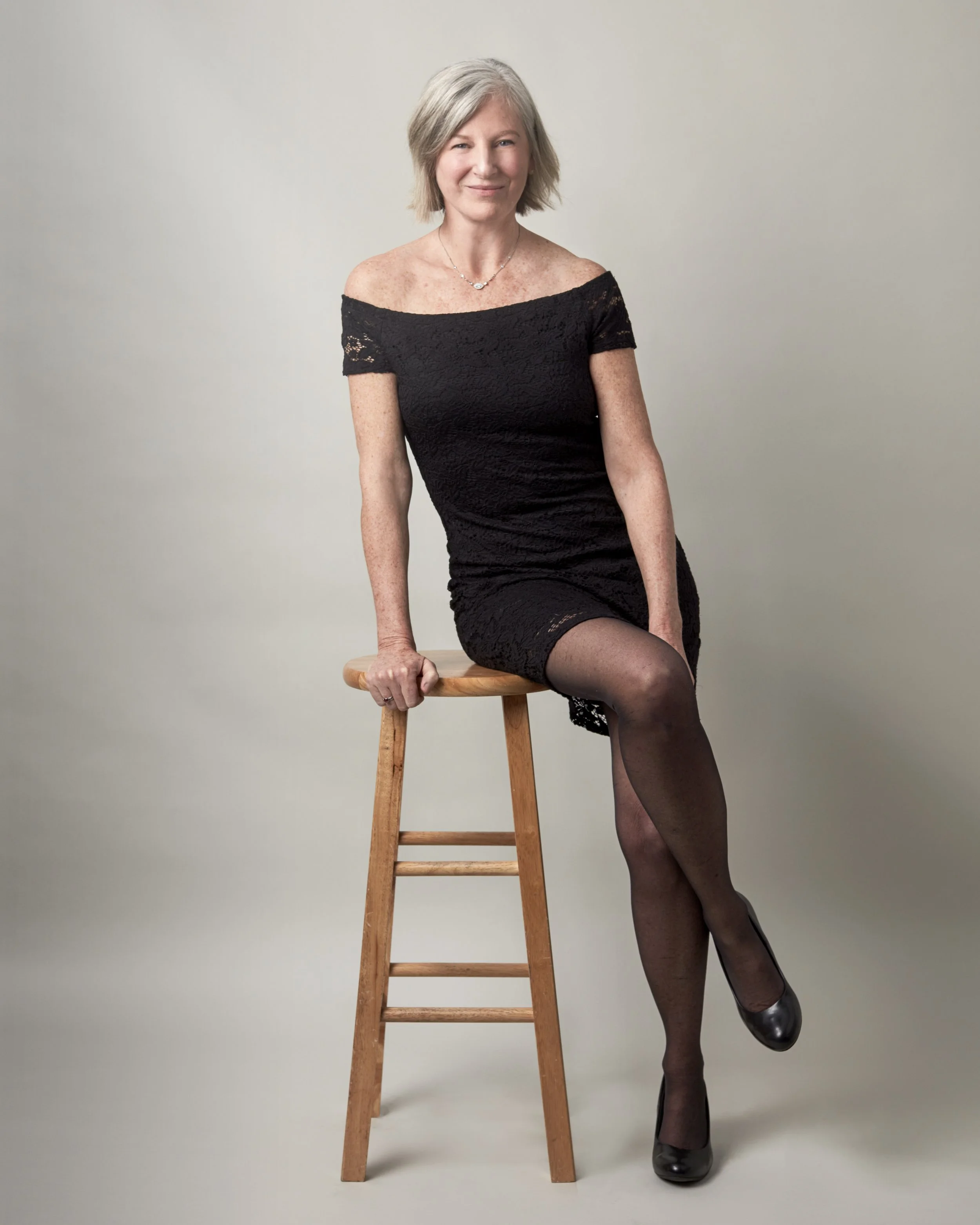 A woman with gray hair sitting on a wooden stool, wearing a black off-the-shoulder lace dress, black stockings, and black heels, posing with one leg crossed over the other against a plain gray background.