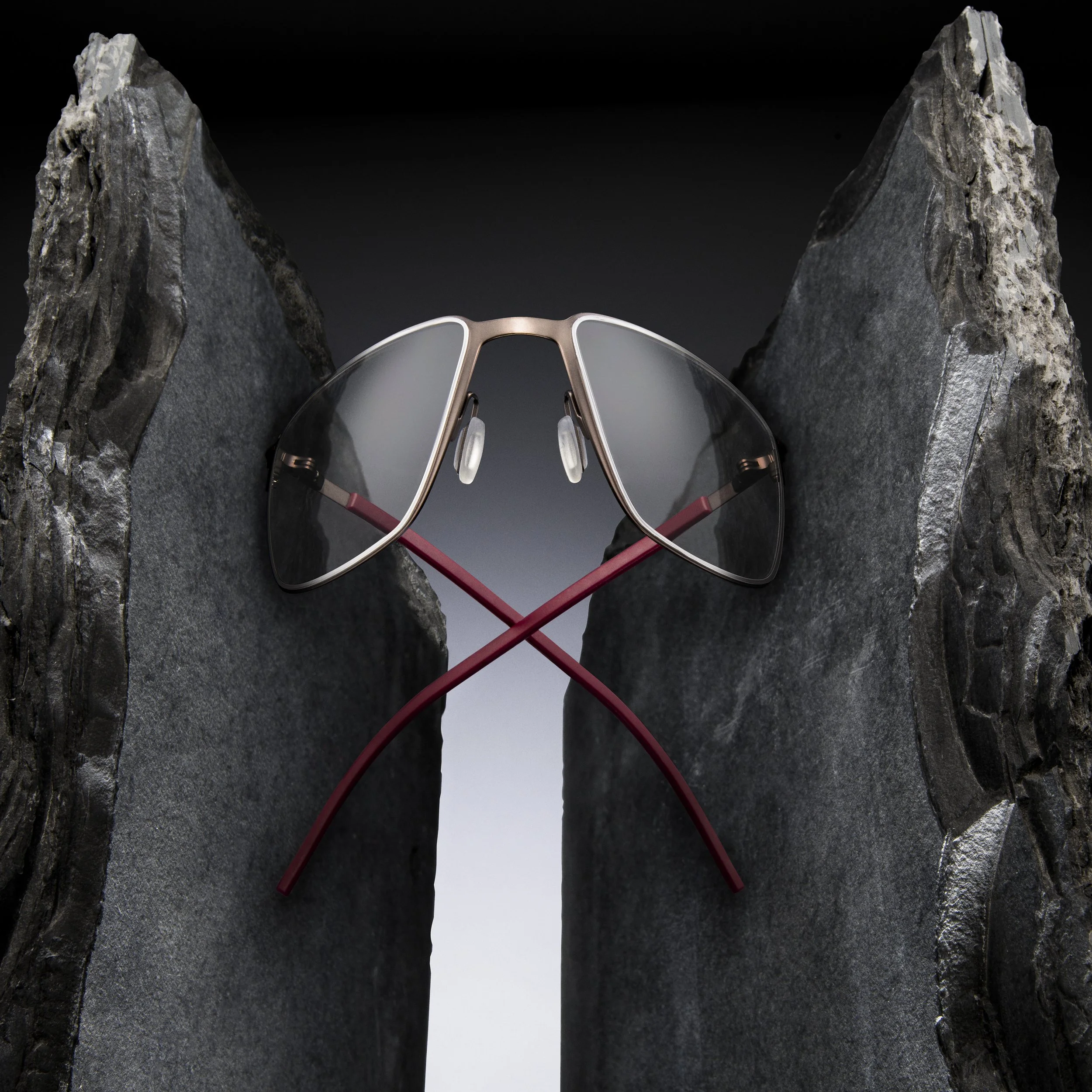 Pair of sunglasses with gray lenses and gold frames resting between two jagged black rocks against a dark background.