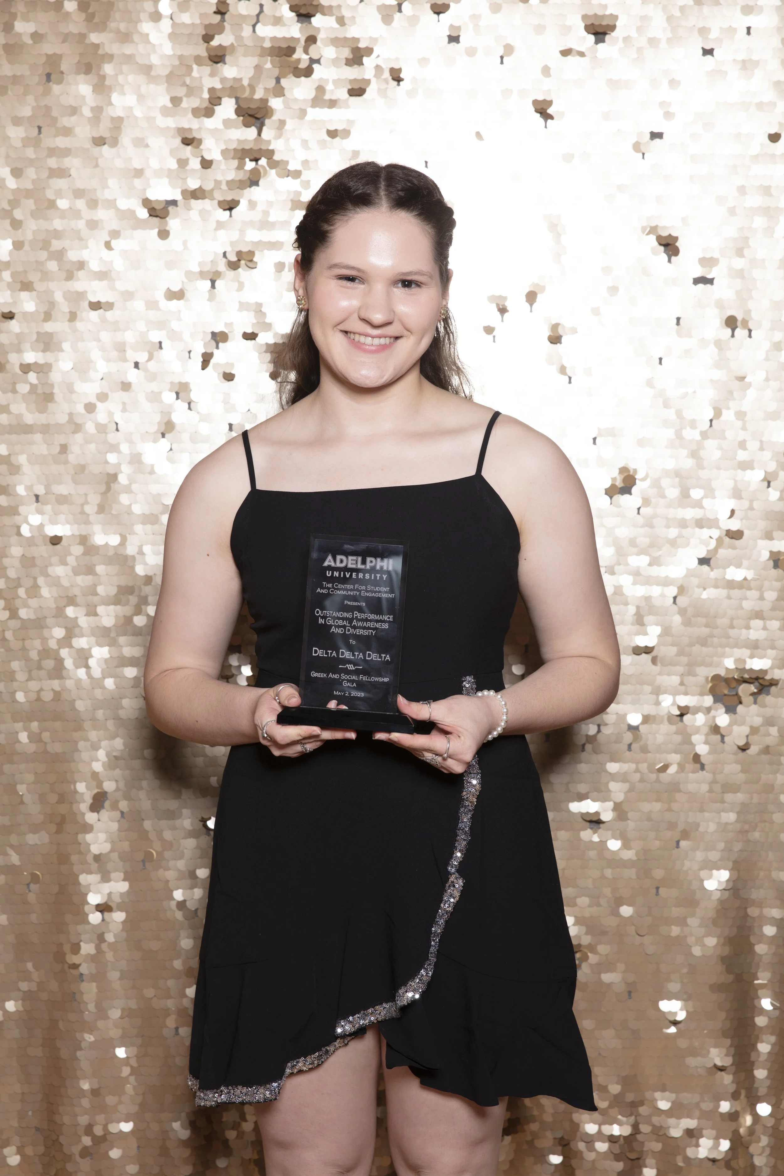 A young woman in a black dress holding an award plaque, standing in front of a gold sequin backdrop.